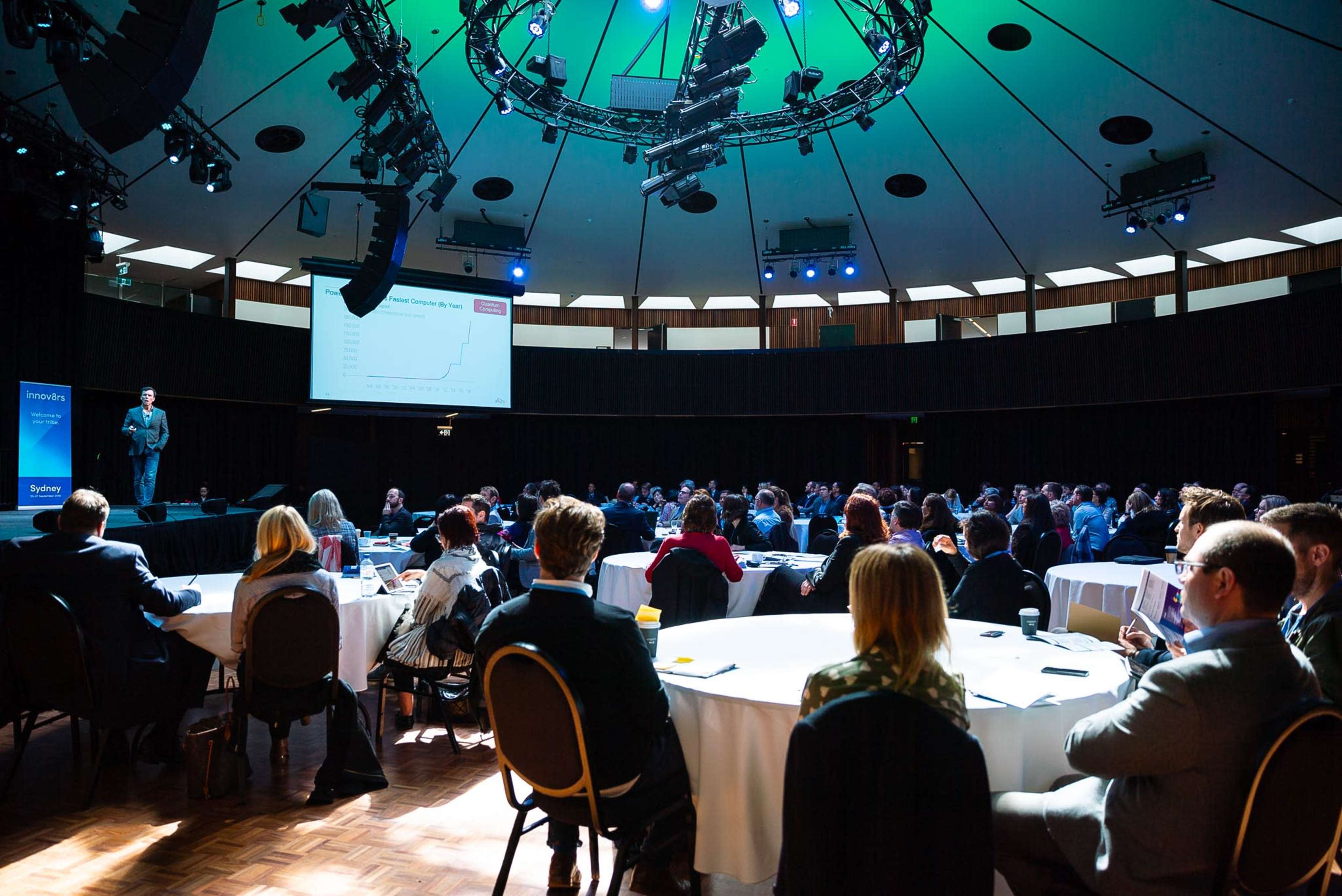 A speaker presents to an audience in a large, round room with circular lighting fixtures and a screen displaying a graph.
