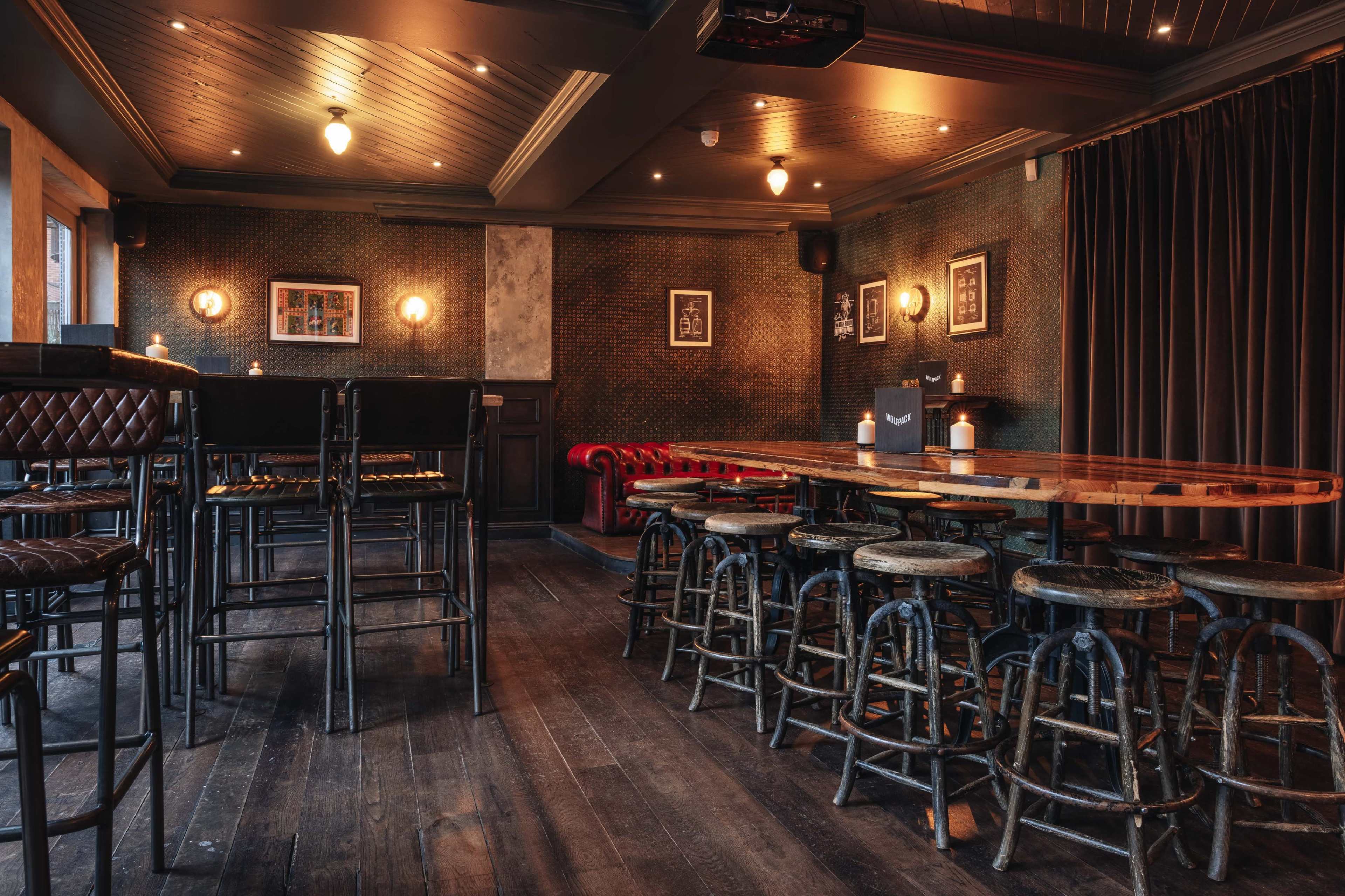 The image shows a dimly lit bar interior with wooden floors, tall and low seating arrangements, and vintage-style decor.