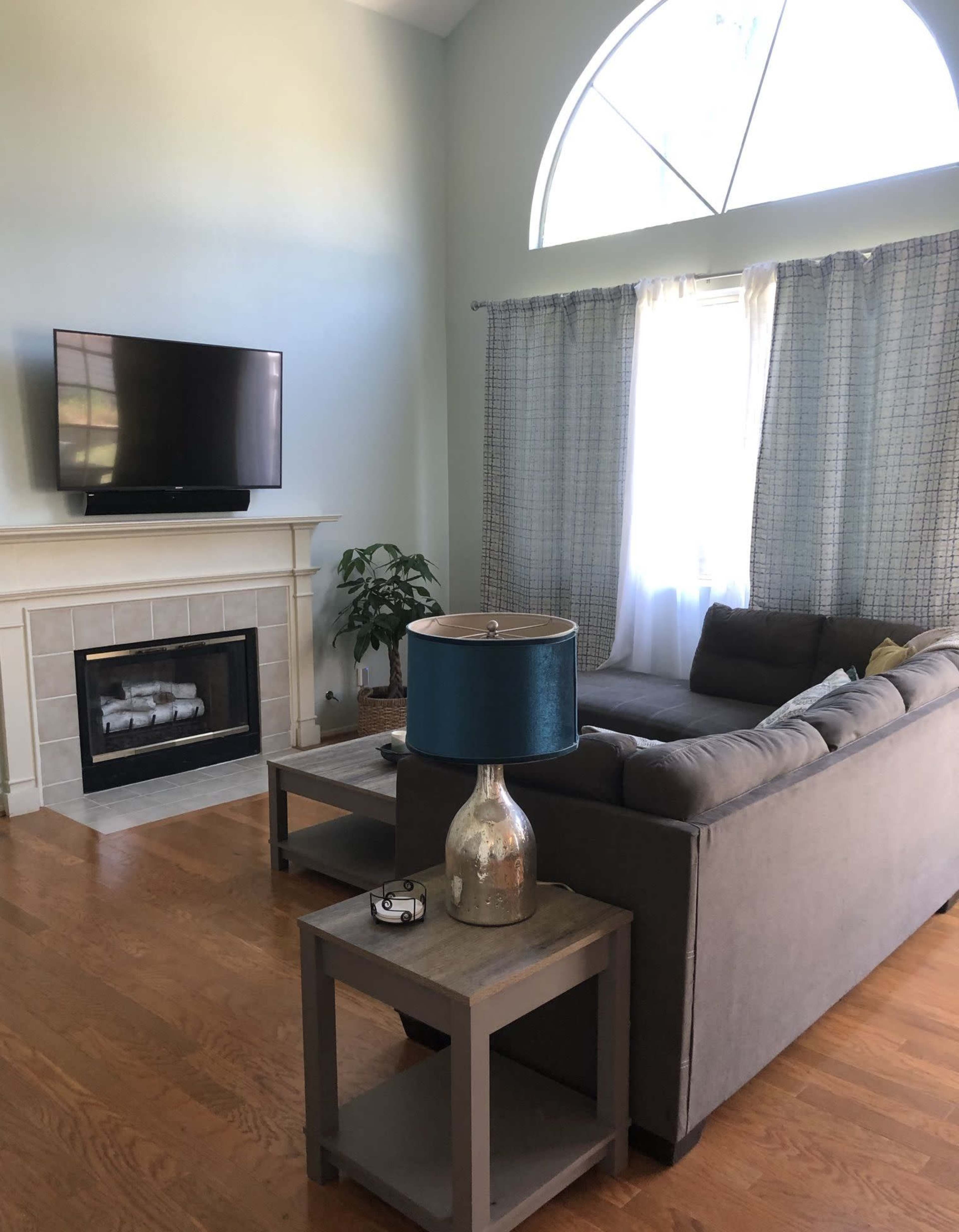 A living room features a gray sofa, a glass-topped side table with a blue lamp, a fireplace, and a mounted television above the mantel.