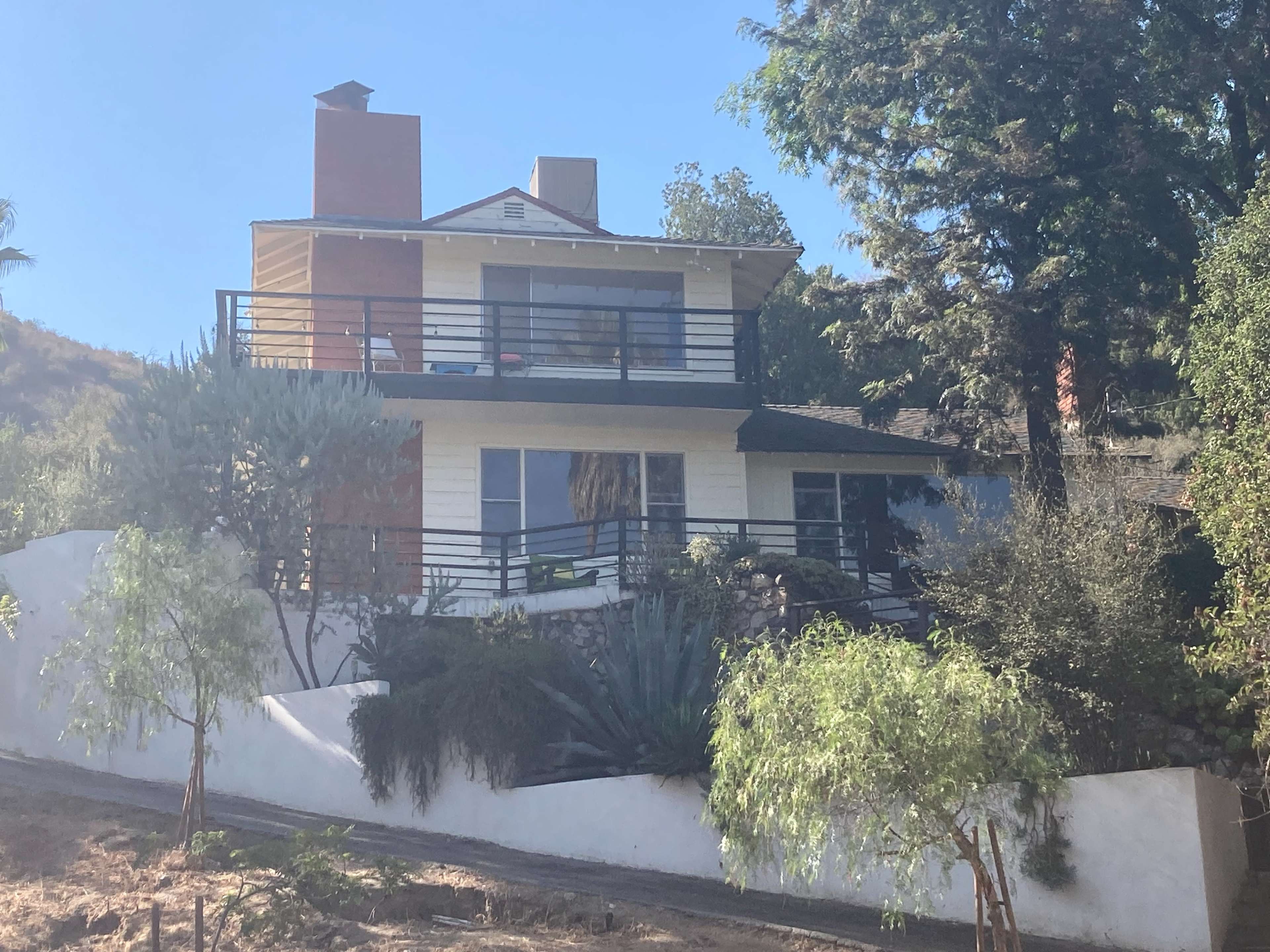 A two-story house with a balcony and chimney is situated on a sloped lot, surrounded by trees and shrubs.