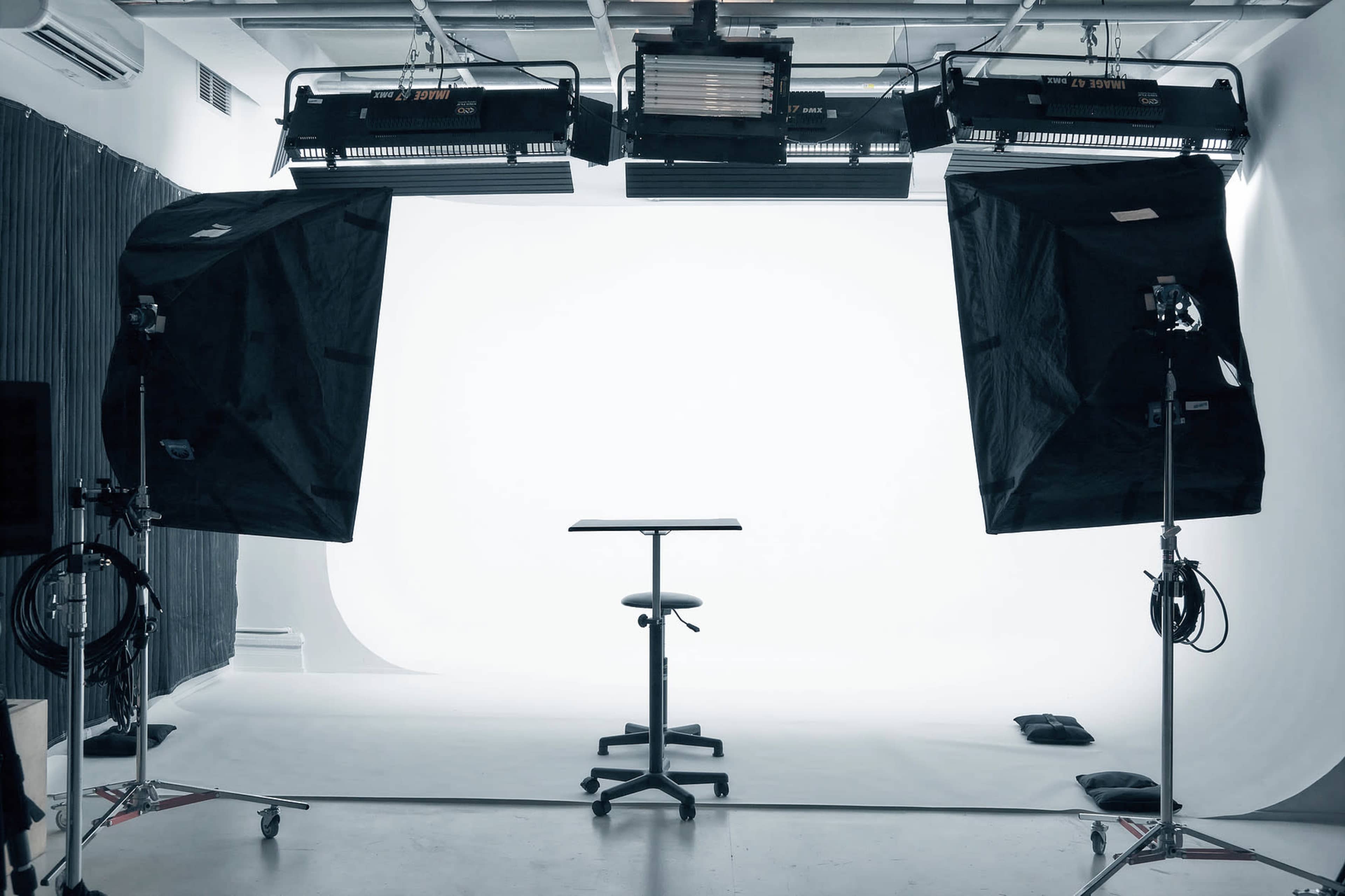 The image shows a photography studio equipped with bright lights and a simple backdrop, featuring a table and a stool in the center.