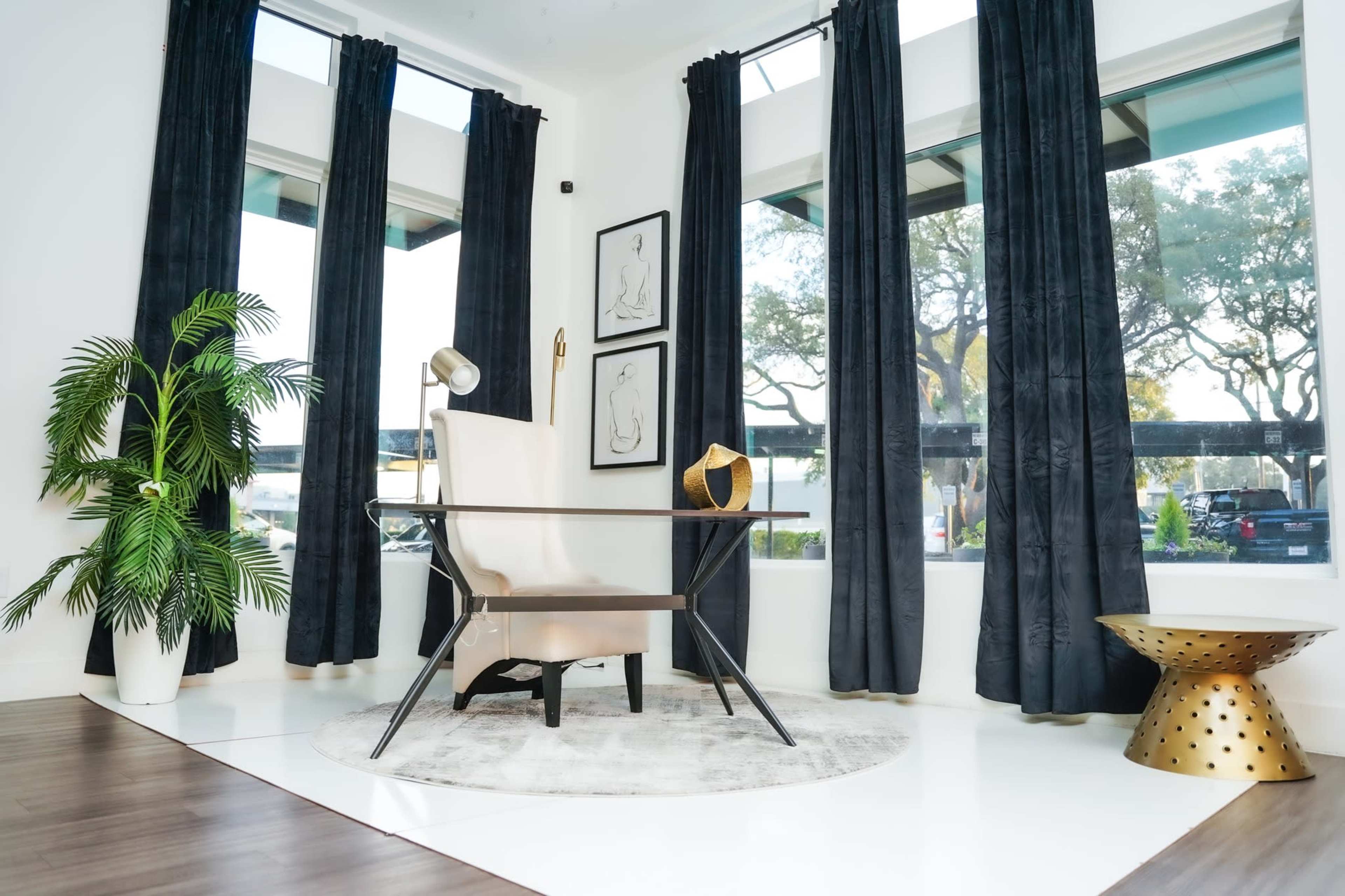 The image shows a modern interior featuring a sleek chair, a small side table, a potted plant, and large windows with dark curtains.