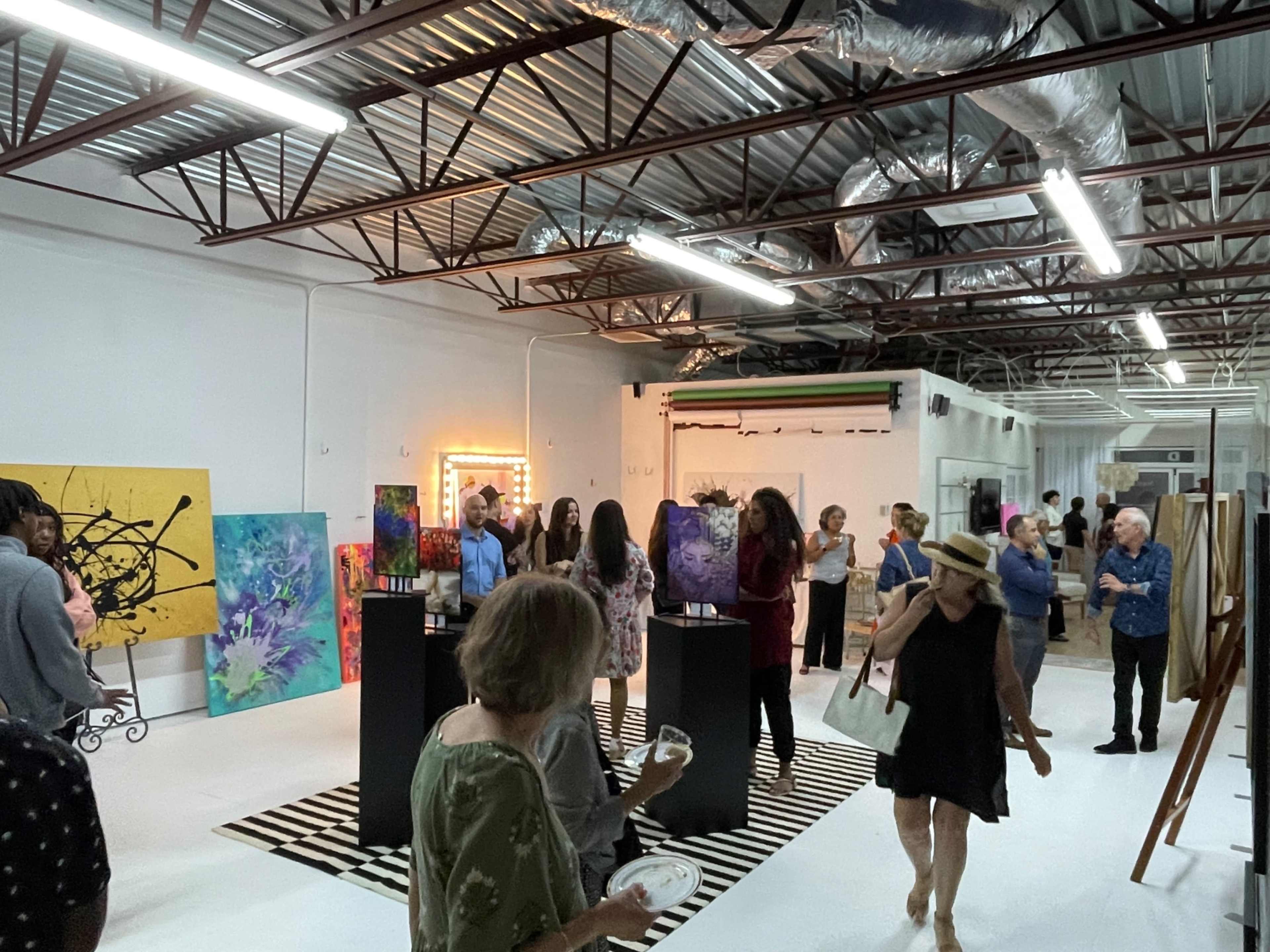 A gallery space filled with people admiring abstract art on display, with several vibrant paintings and a striped rug on the floor.