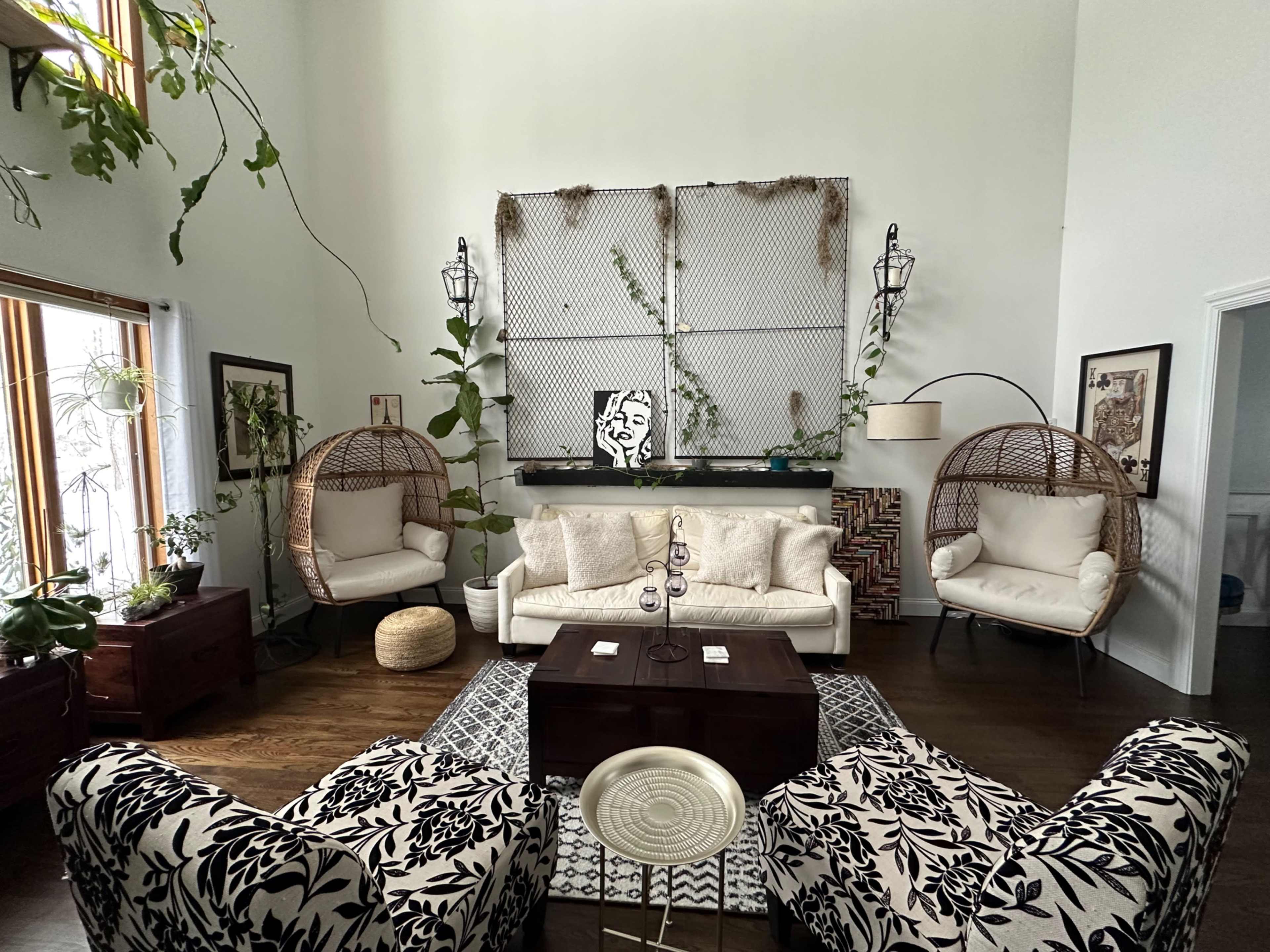 The living room features two wicker chairs, a white sofa, and wall-mounted plants, with a large artwork displayed above a wooden table.
