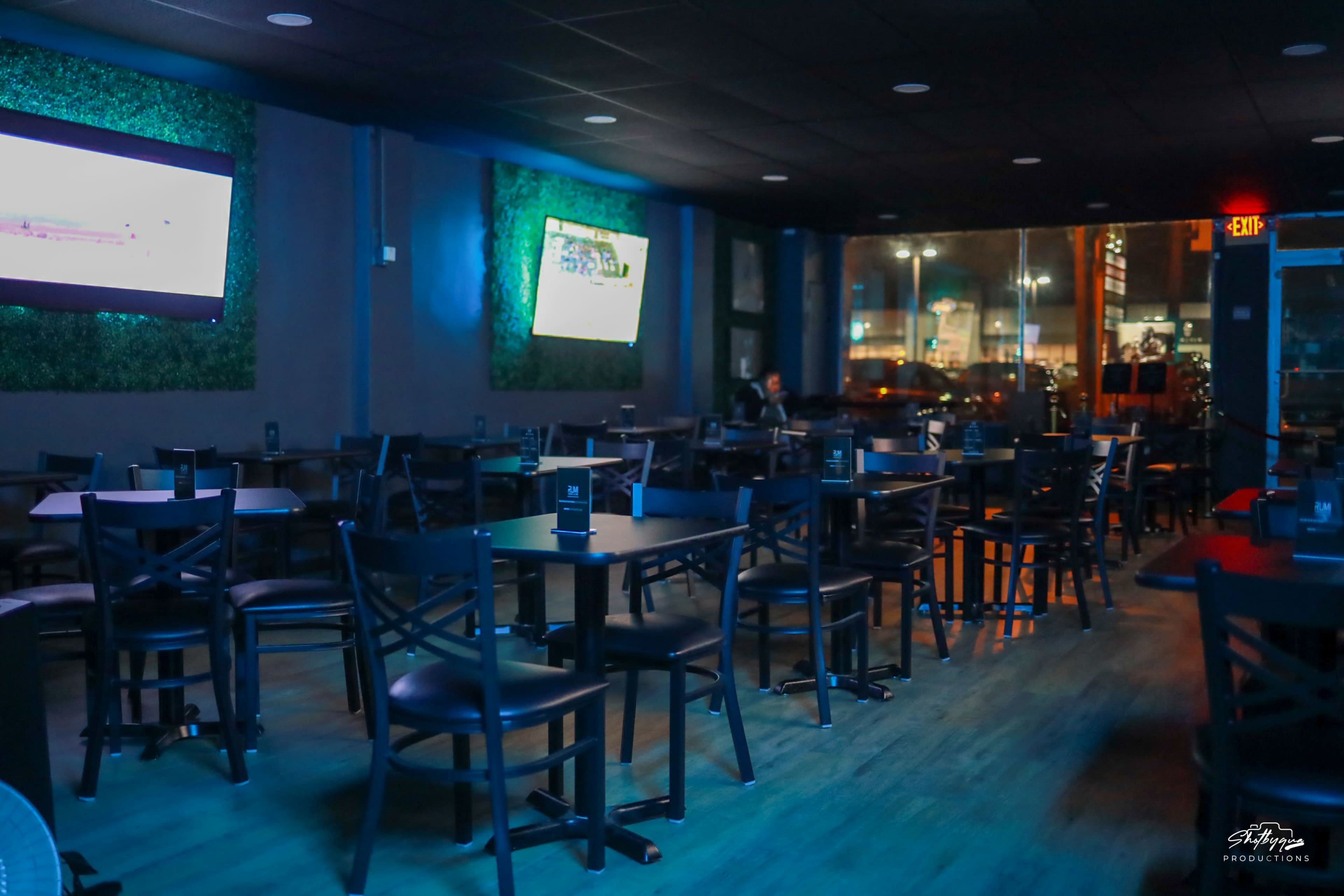An empty bar with black tables and chairs, featuring two televisions on the walls and large windows displaying a city view at night.