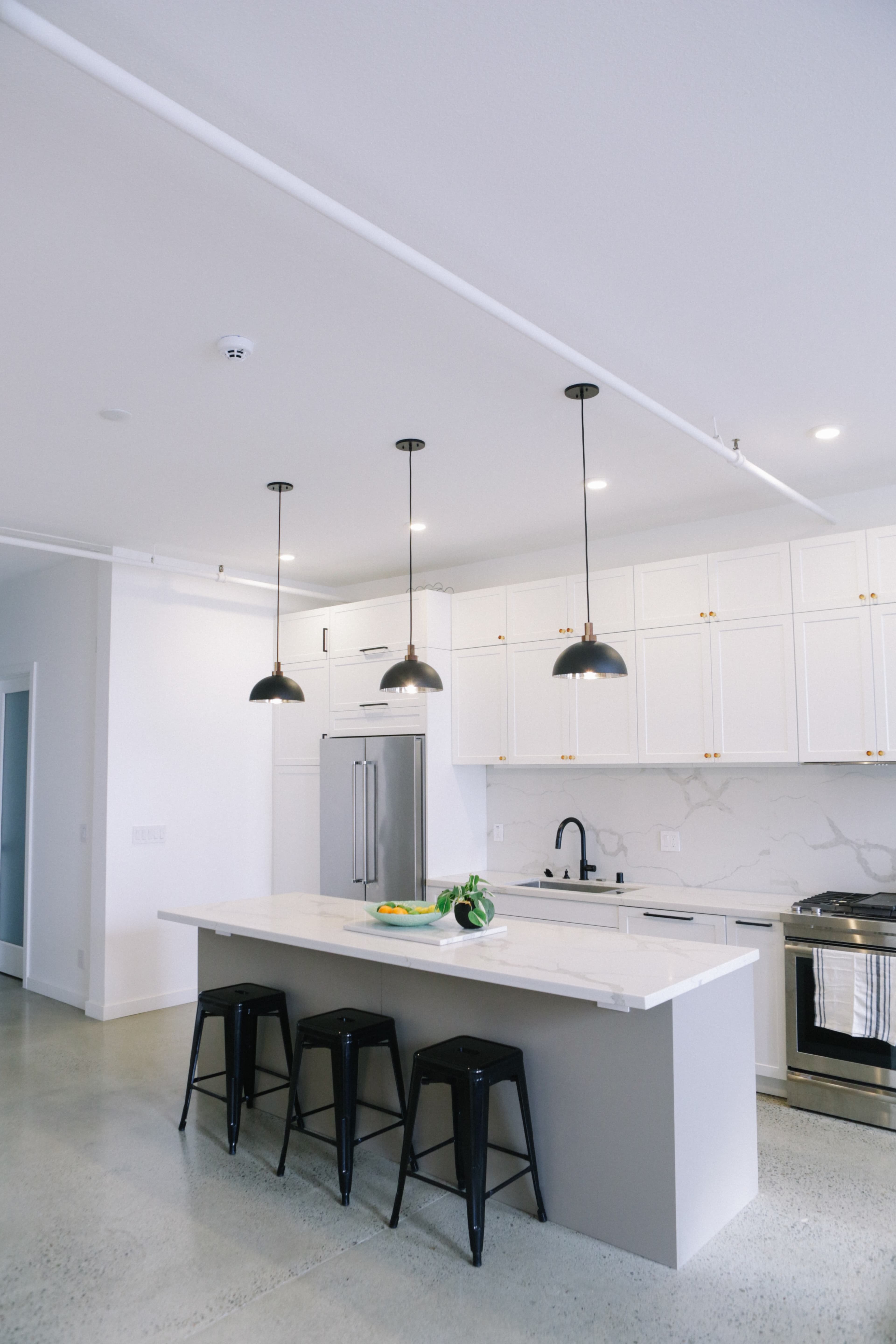 A modern kitchen features a large island with a white countertop, black pendant lights overhead, and minimalistic black stools.
