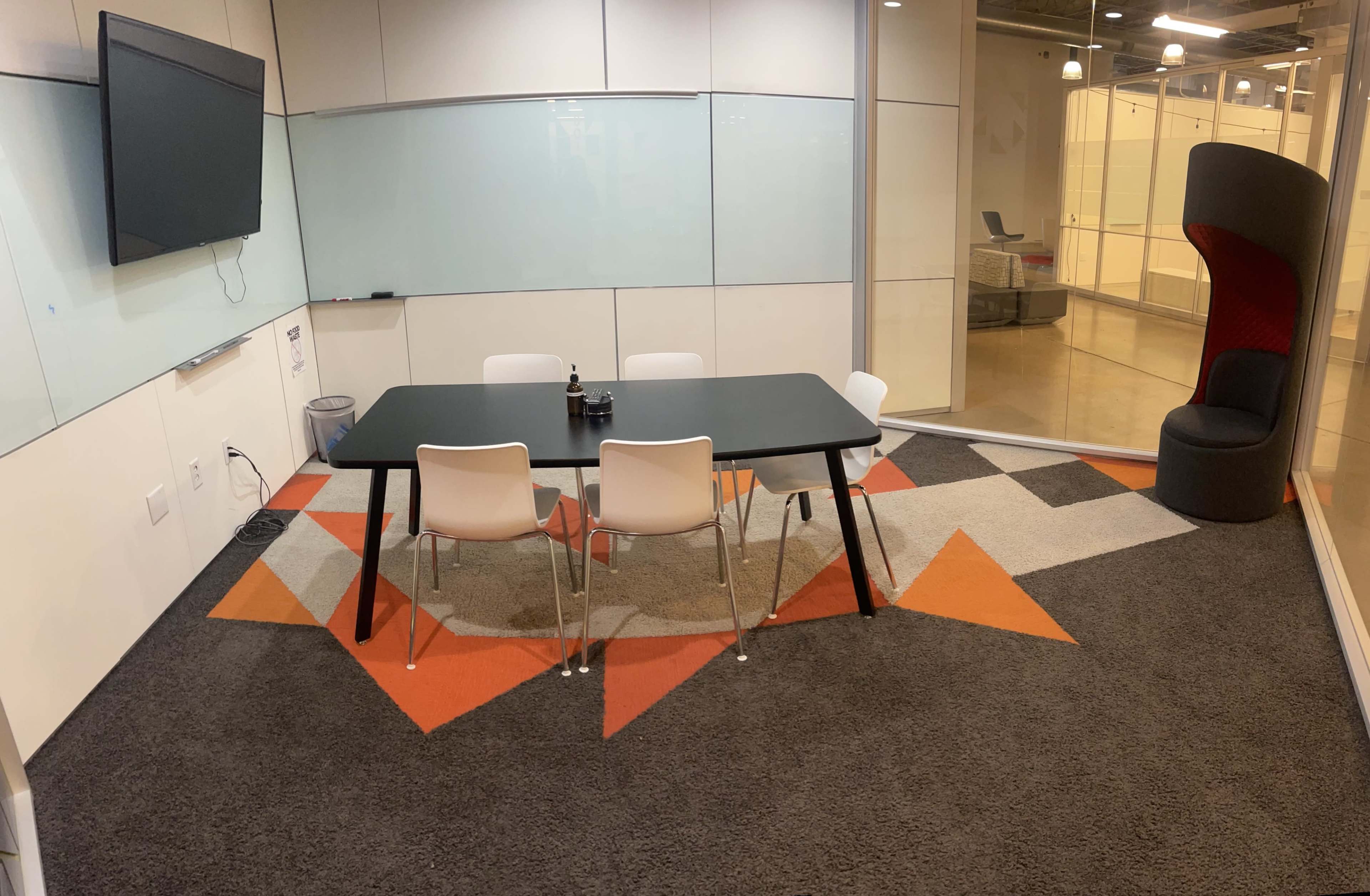 A modern meeting room with a black table surrounded by white chairs on a patterned orange and gray carpet, featuring a wall-mounted TV and a separate red acoustic chair in one corner.