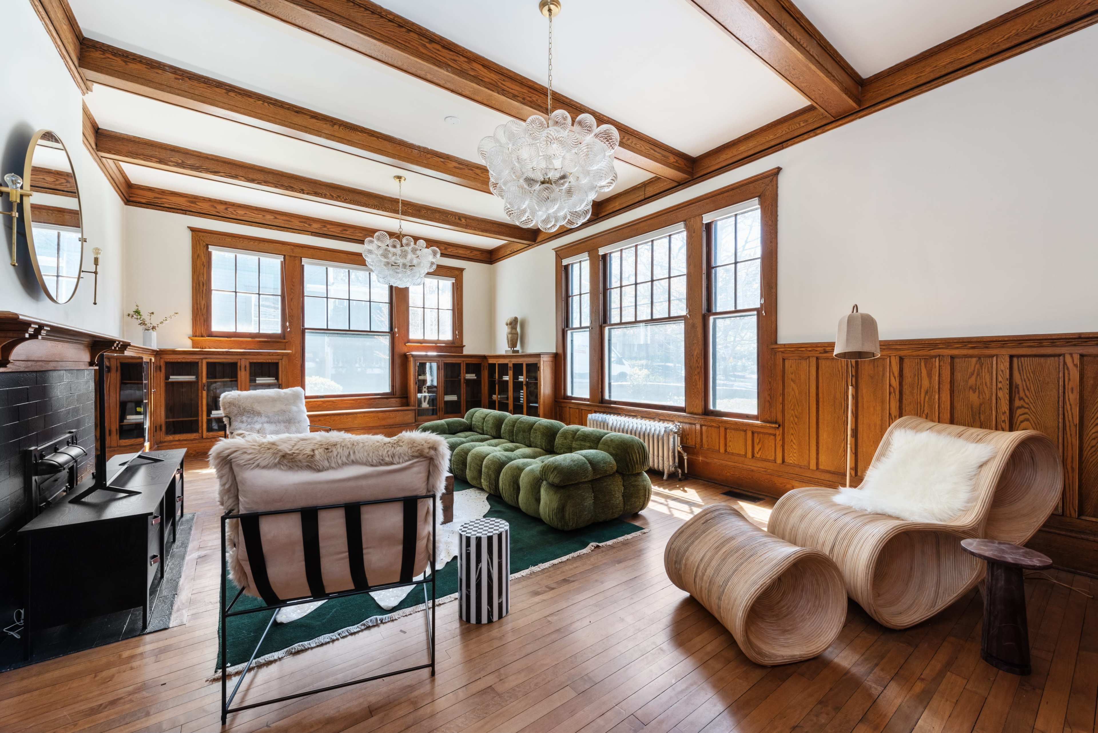 The image depicts a spacious living room featuring wooden beams, large windows, a green sectional sofa, and various modern furniture pieces.