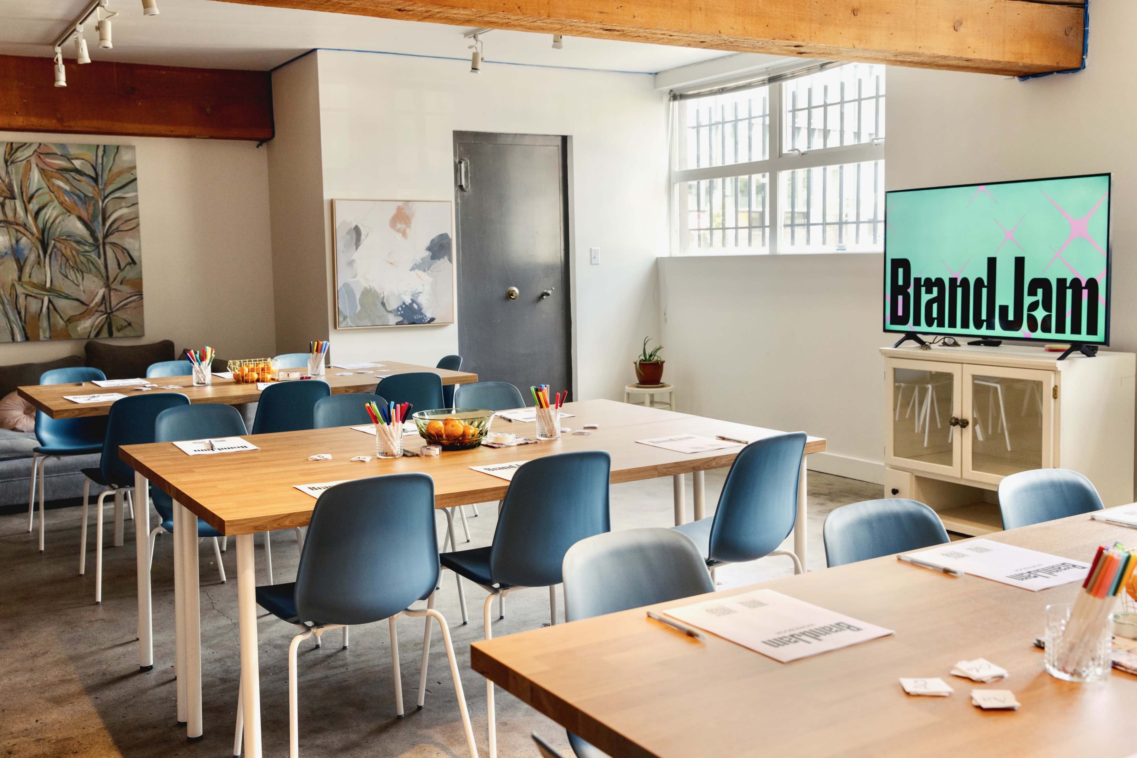 A spacious workshop room features several wooden tables with chairs, colorful writing utensils, and a large screen displaying "BrandJam."