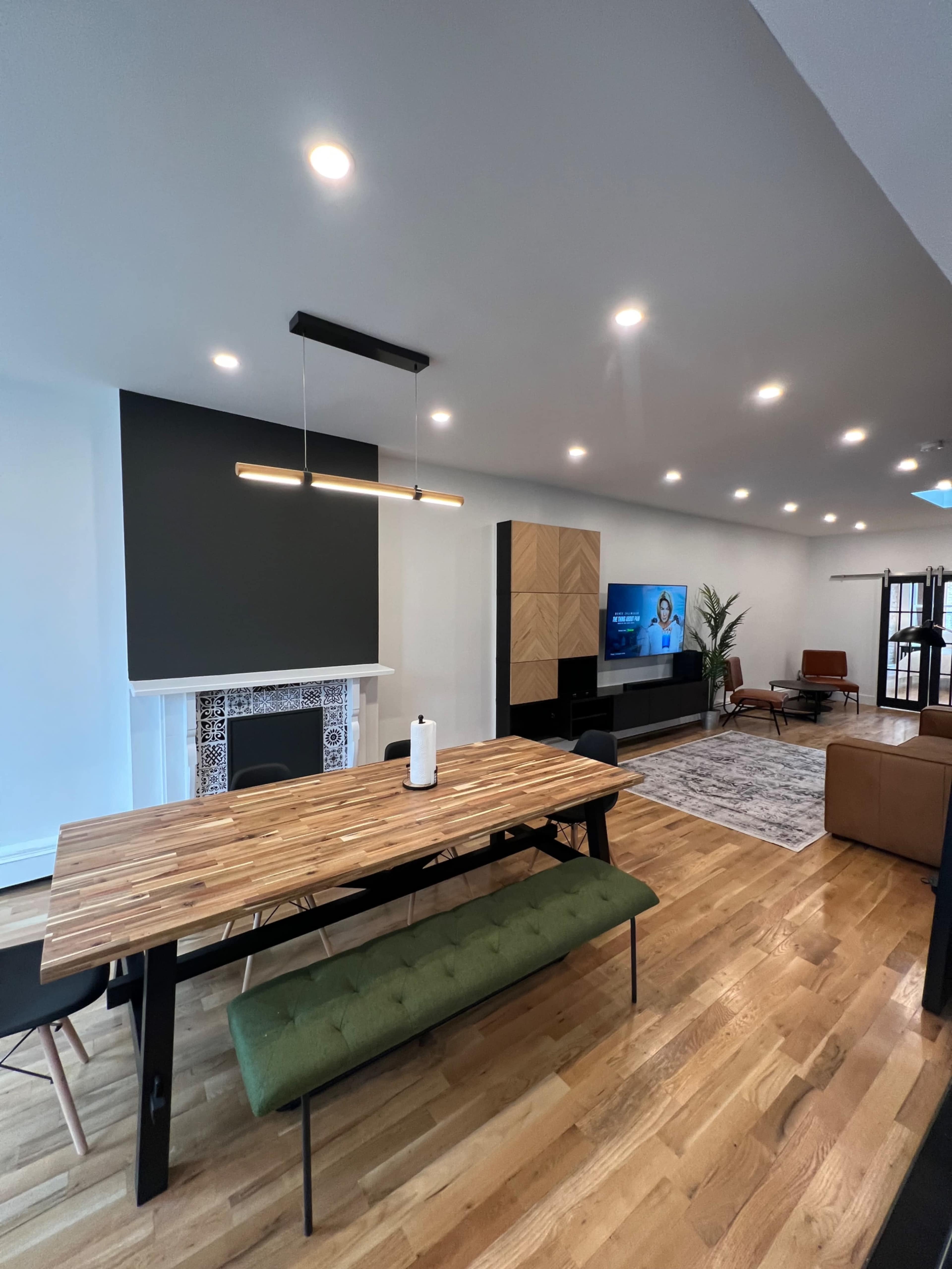 A modern living and dining area features a wooden dining table with a green bench, a TV mounted on the wall, and a cozy seating arrangement.