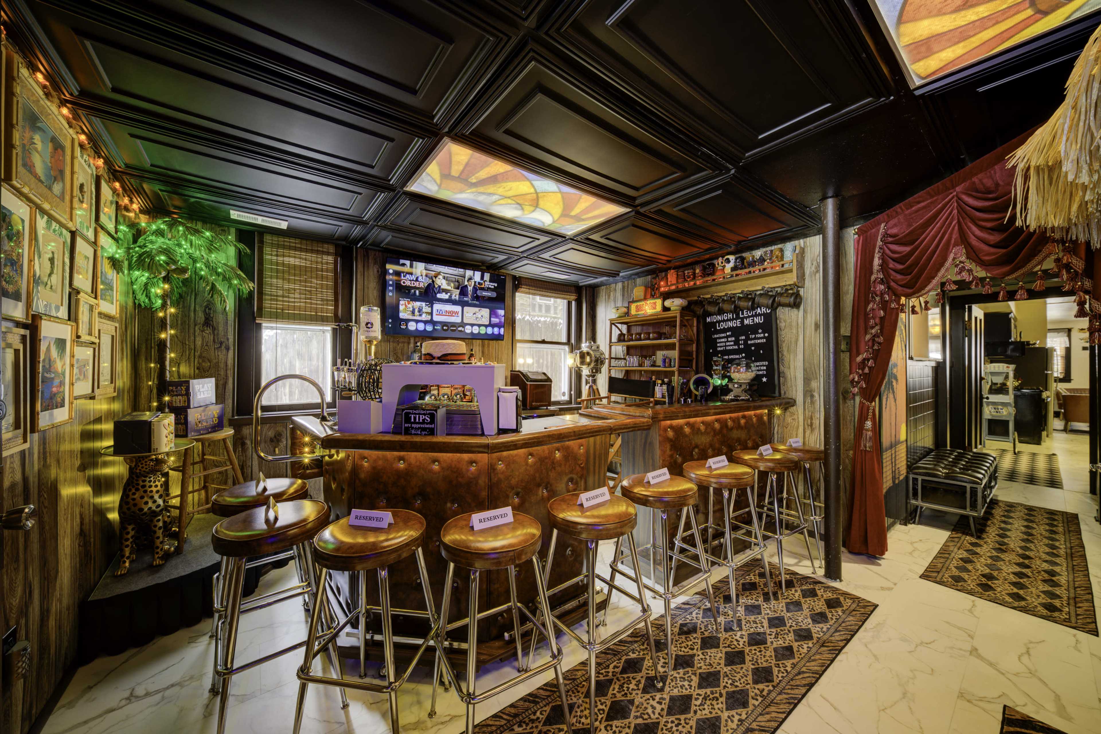 The image shows a warmly lit bar area featuring a dark wood ceiling, colorful wall art, and a row of high stools along the counter.