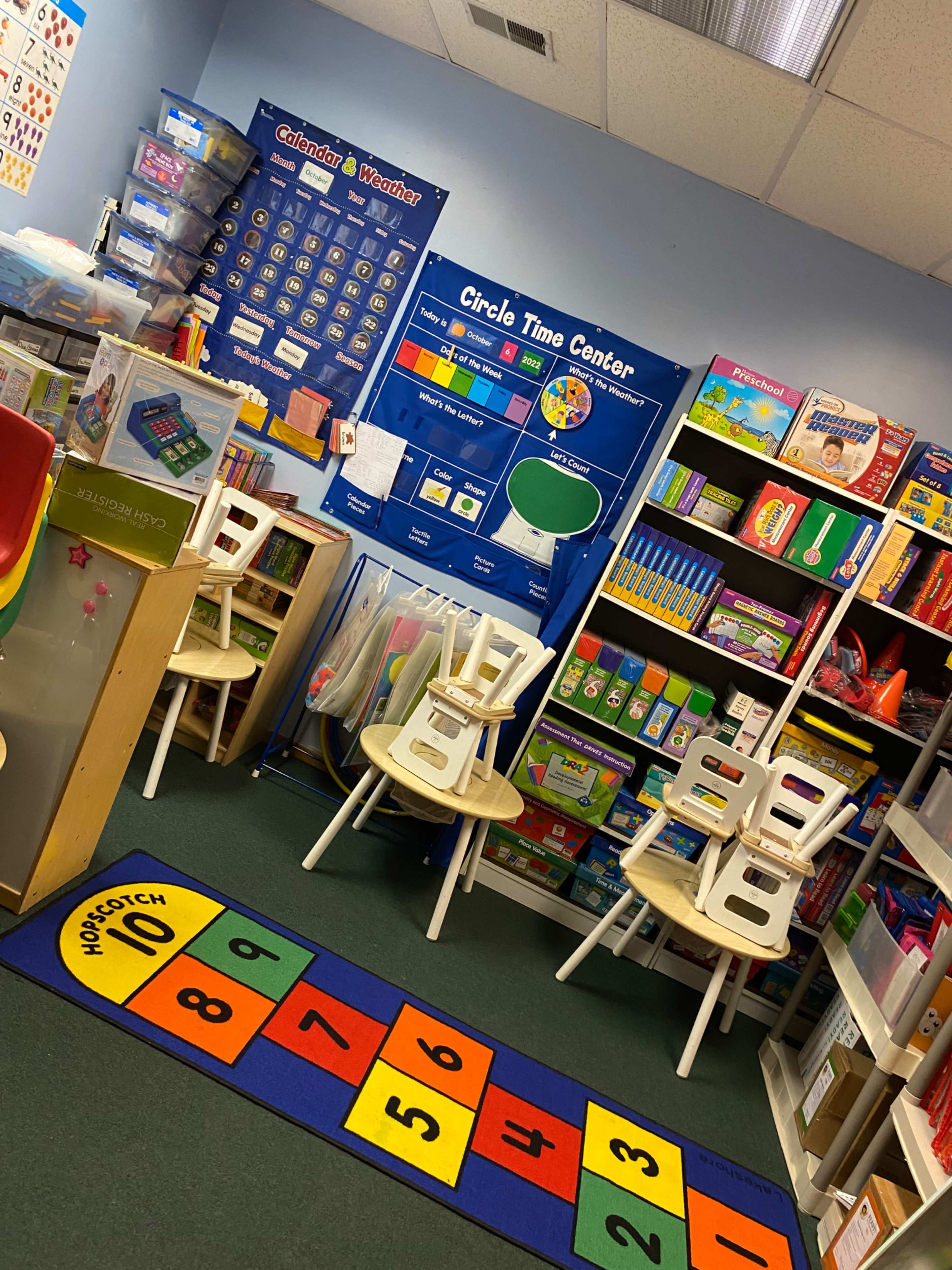 A classroom is shown with colorful educational materials, a calendar, and several stacked chairs surrounding a rug with numbers.