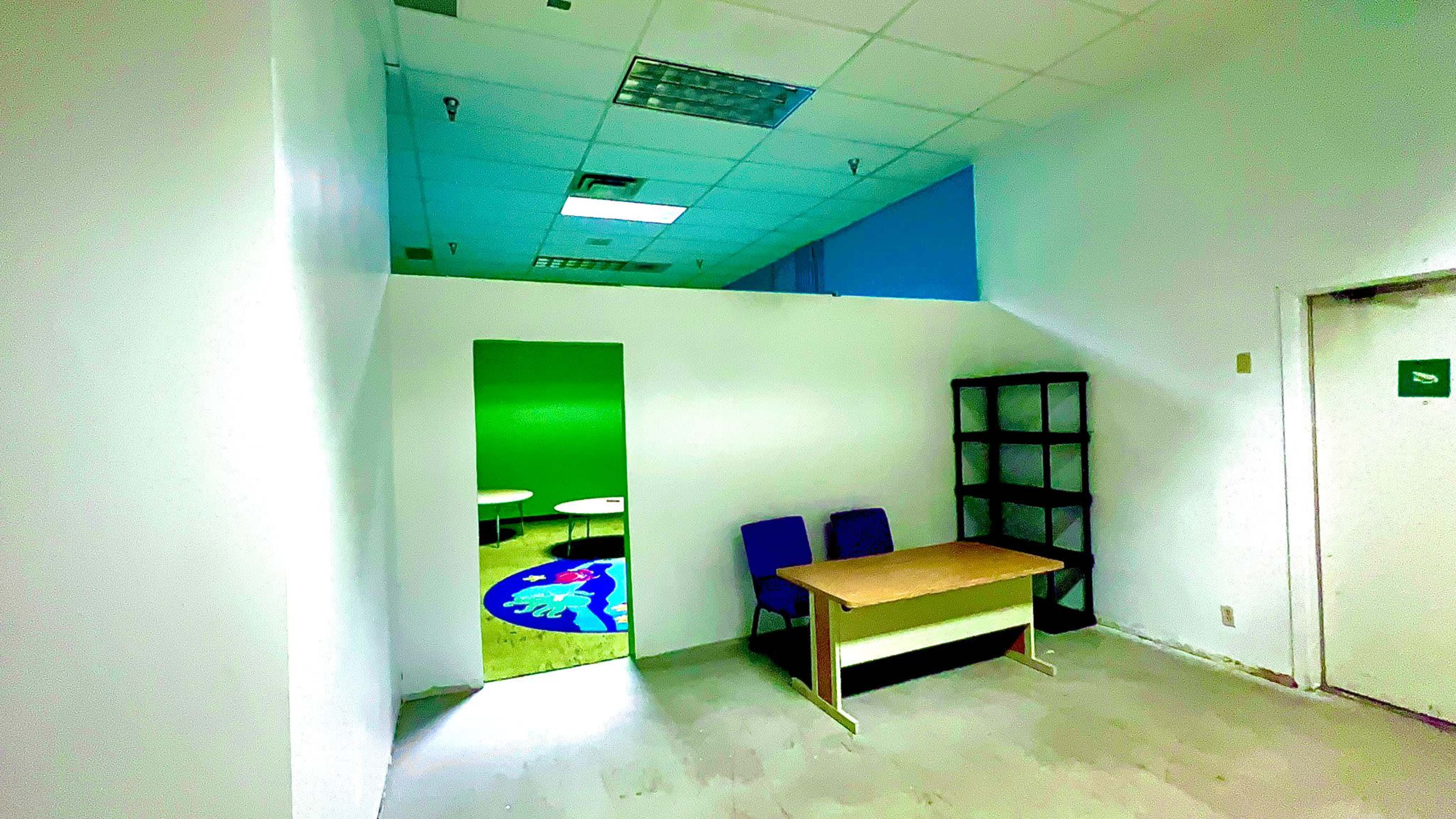 The image shows a sparse office space with a table, two chairs, a bookshelf, and a doorway leading to a green-walled area.