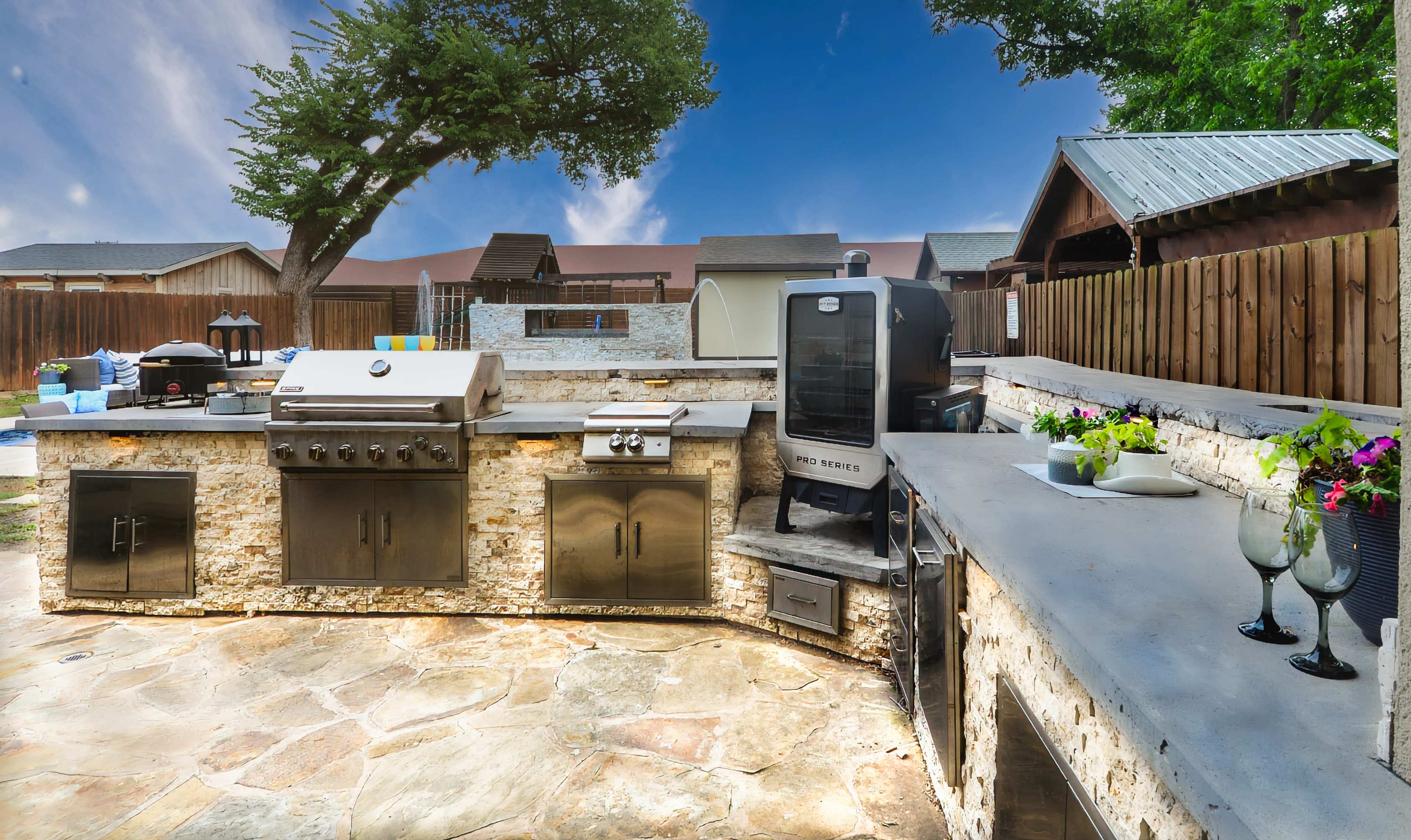 The image shows an outdoor kitchen area featuring a stone countertop, stainless steel appliances, and a grill, surrounded by a landscaped backyard.