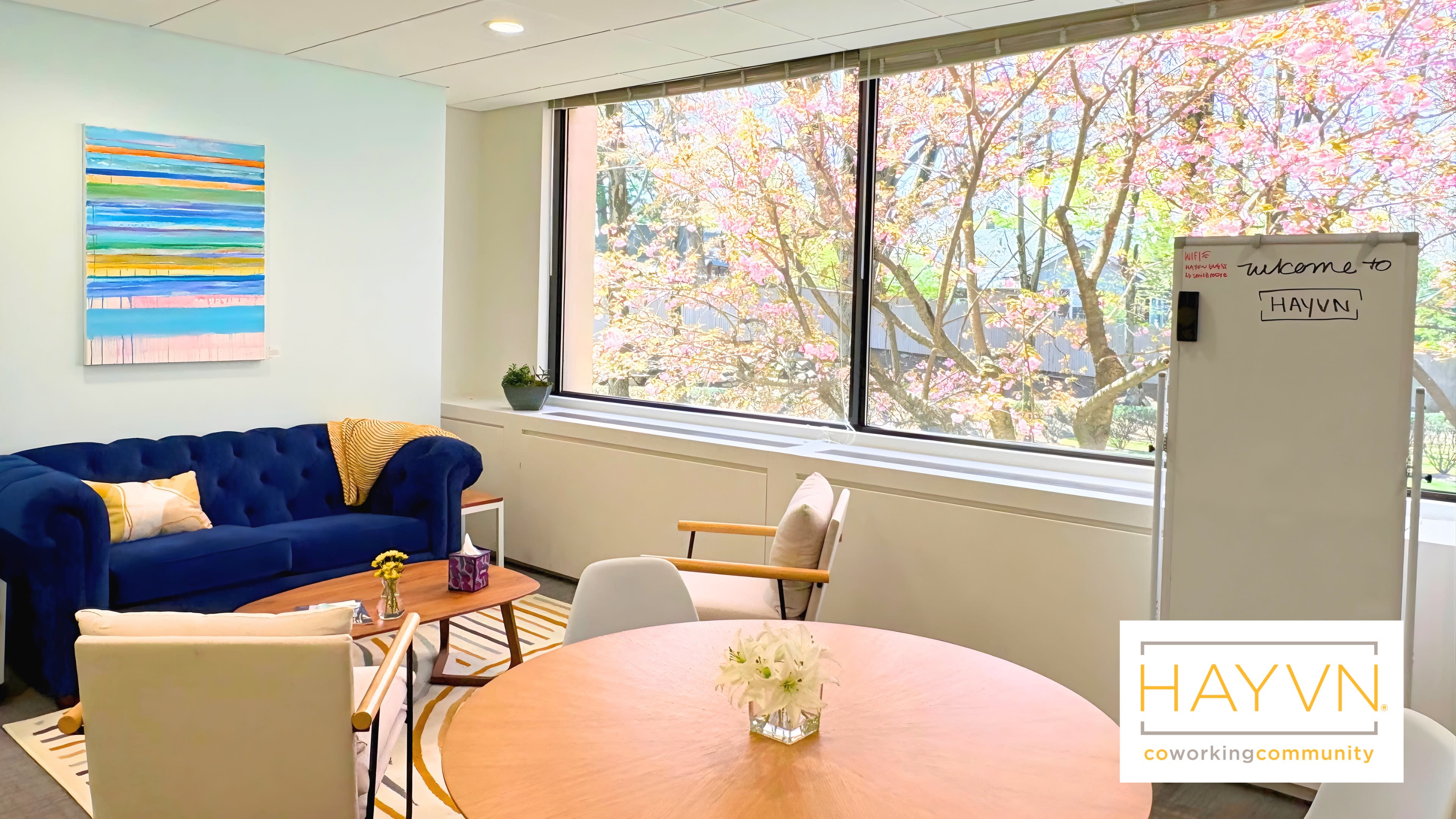 The image shows a bright and modern coworking space featuring a blue sofa, a round table with chairs, and a large window overlooking blooming trees.