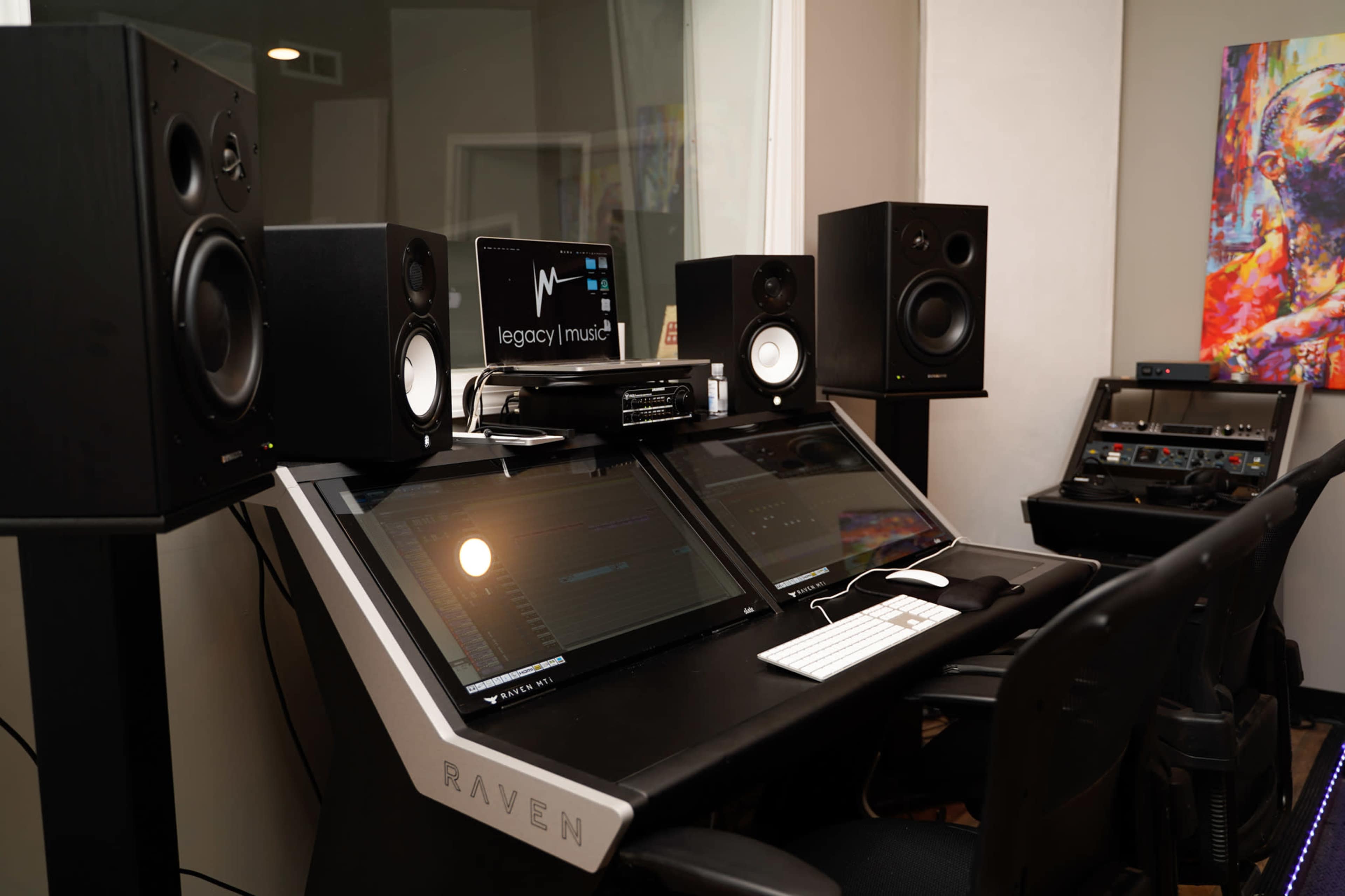 A music production studio featuring a desk with two large monitor screens, audio speakers on either side, and a keyboard, set against a backdrop of colorful artwork.