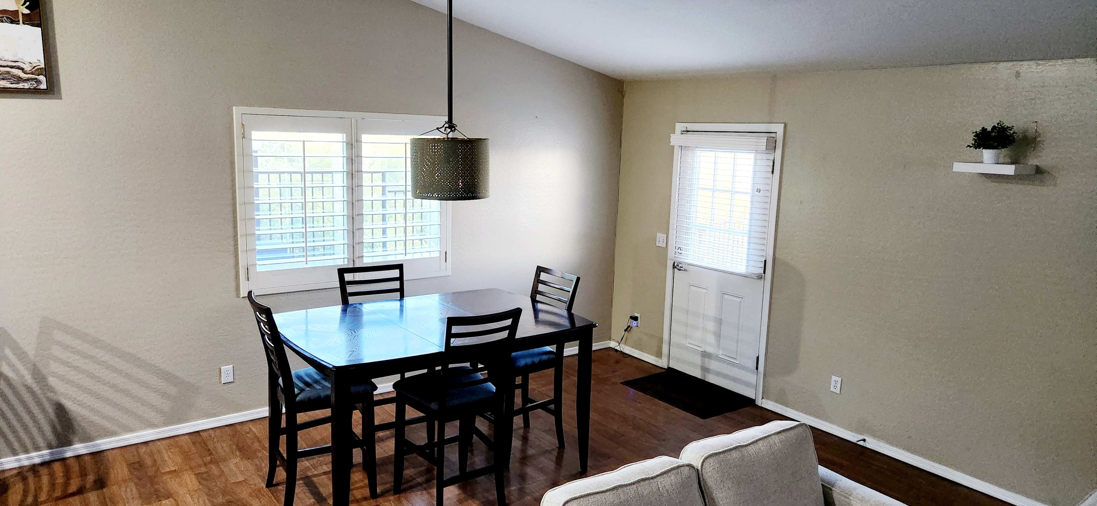 The image shows a small dining area with a black dining table and chairs, a light fixture above, and a door leading outside, all set against a light-colored wall.