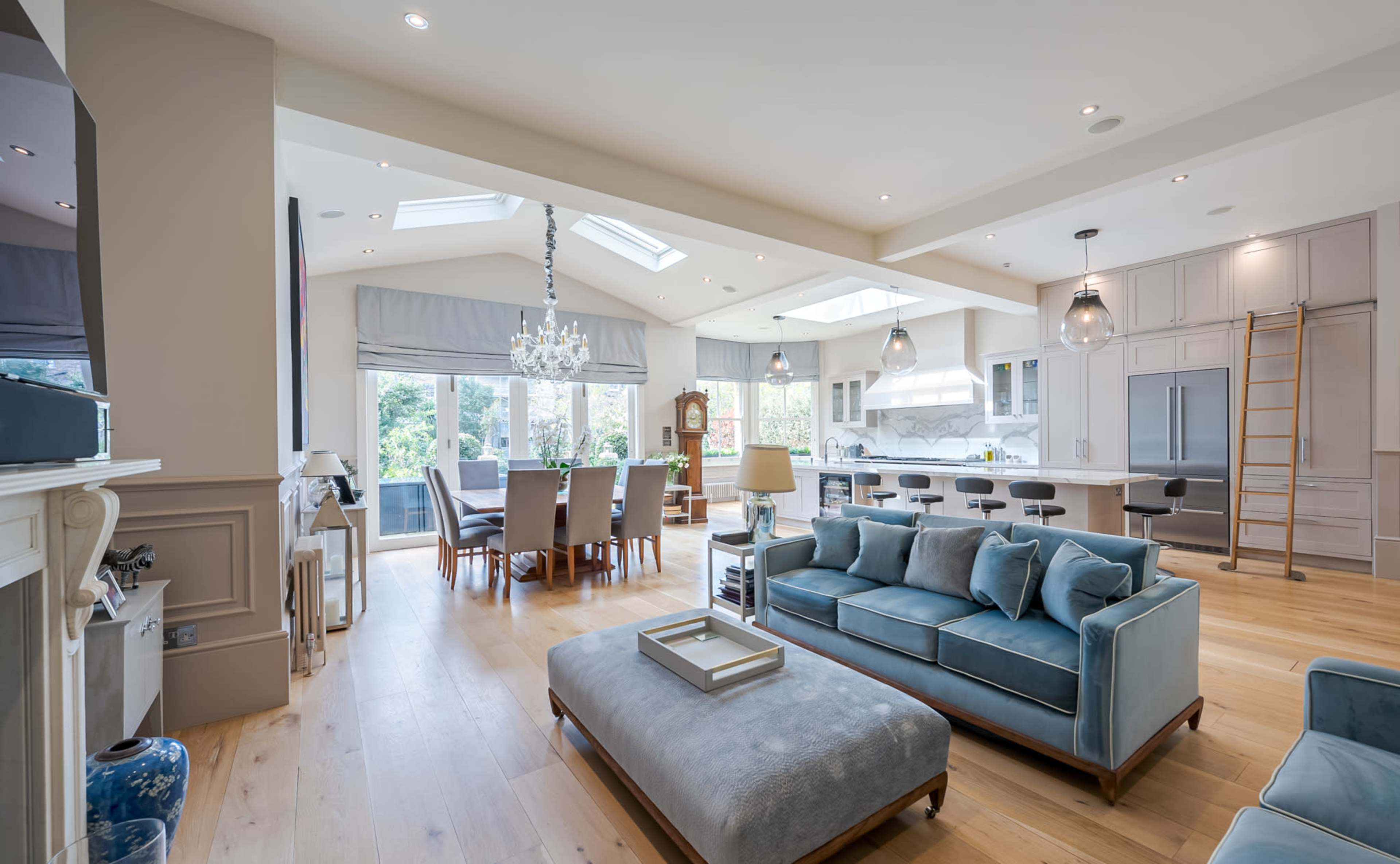 The image shows a modern open-concept living space featuring a blue sofa, a dining area with a chandelier, and a kitchen with bar stools and large windows.