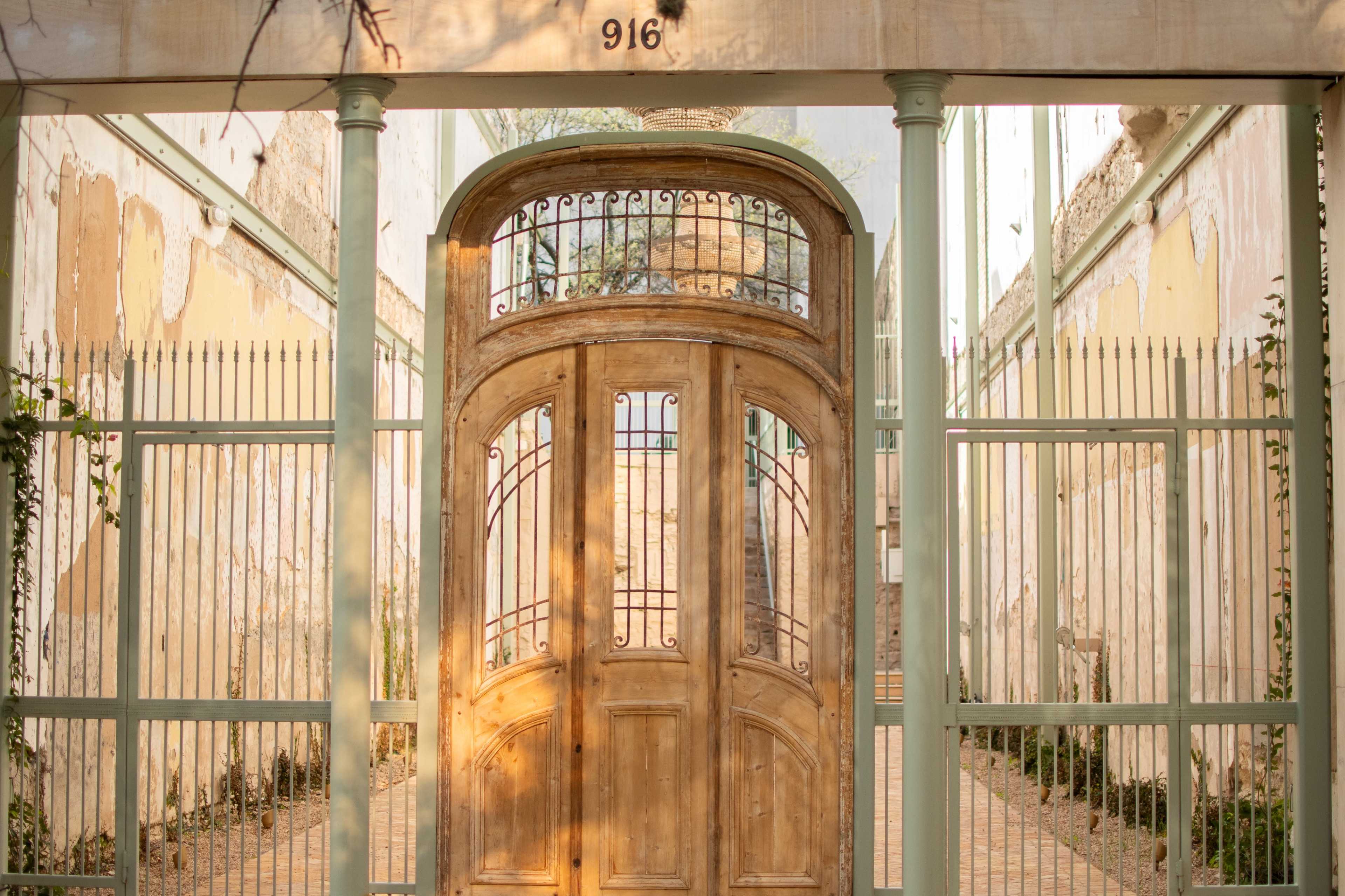 The image shows an intricately designed wooden gate with wrought iron accents, set within a weathered stone enclosure and flanked by pillars.