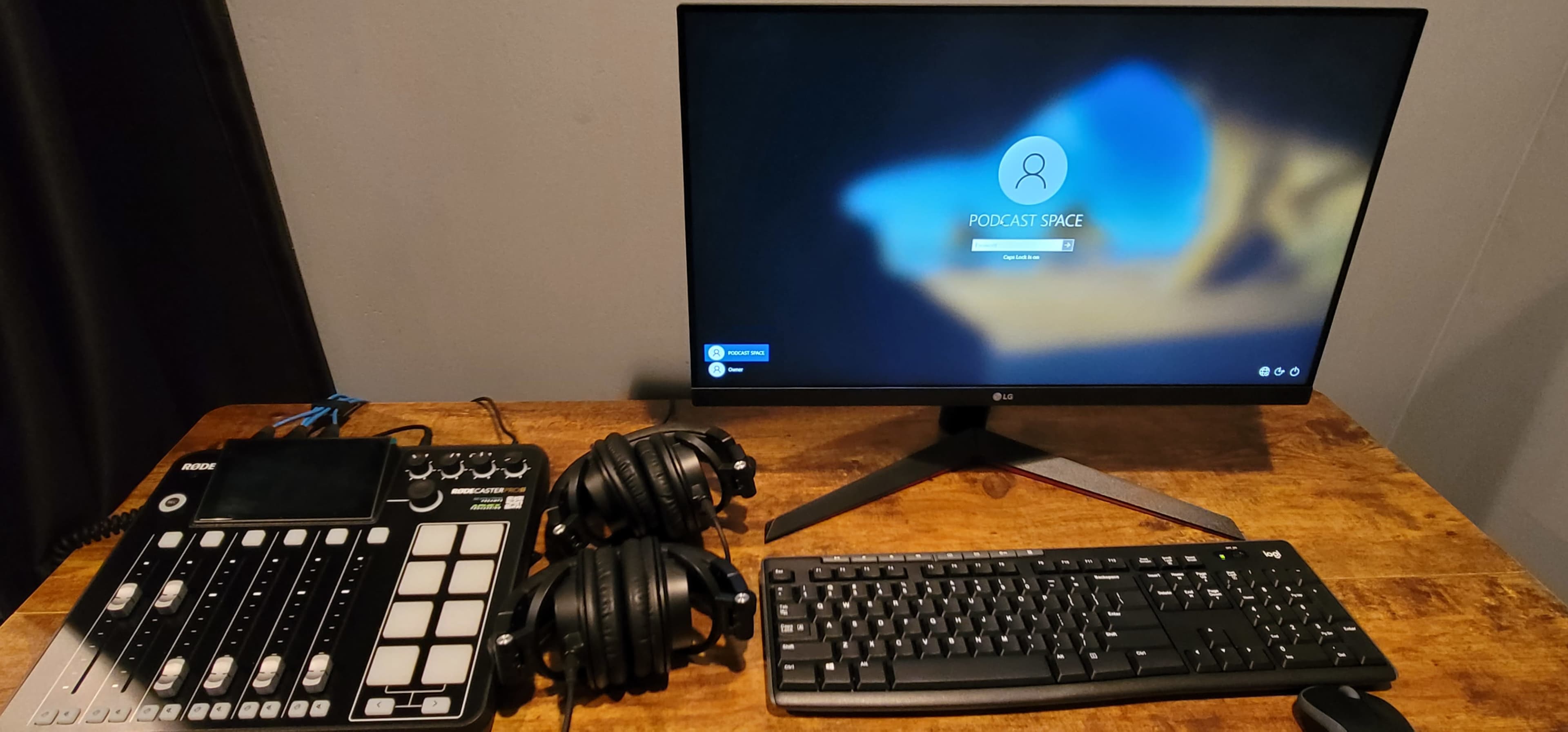 A podcasting setup featuring a computer monitor displaying a login screen, a digital audio mixer, two sets of headphones, and a keyboard arranged on a wooden desk.