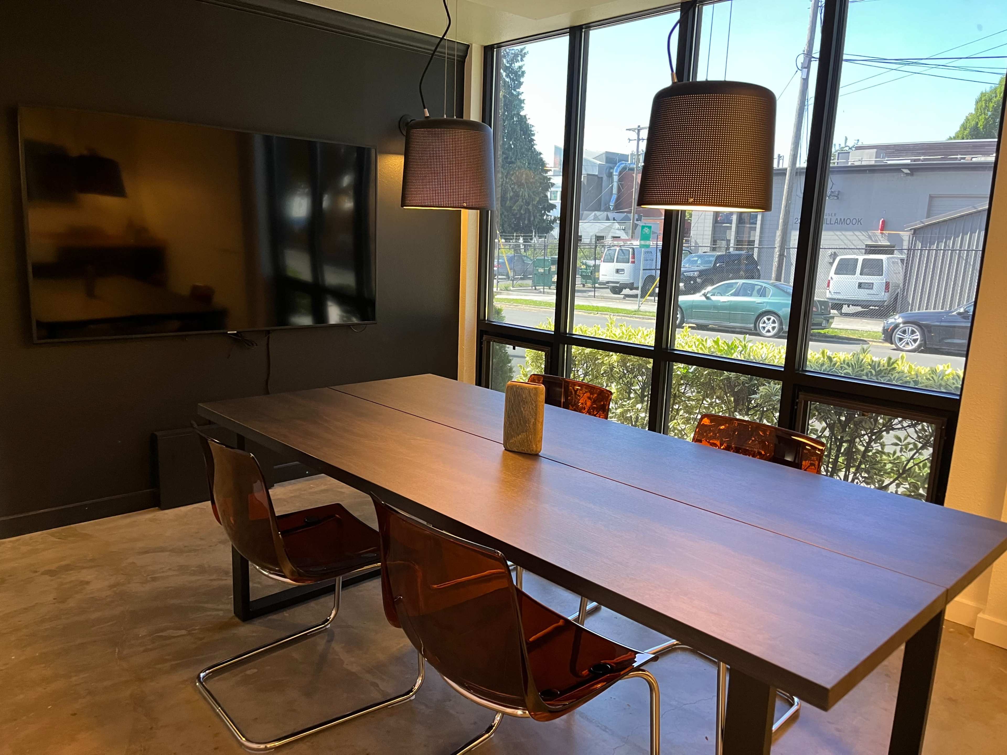 A modern conference room features a long table with four chairs, a television mounted on the wall, and large windows overlooking an outdoor area.