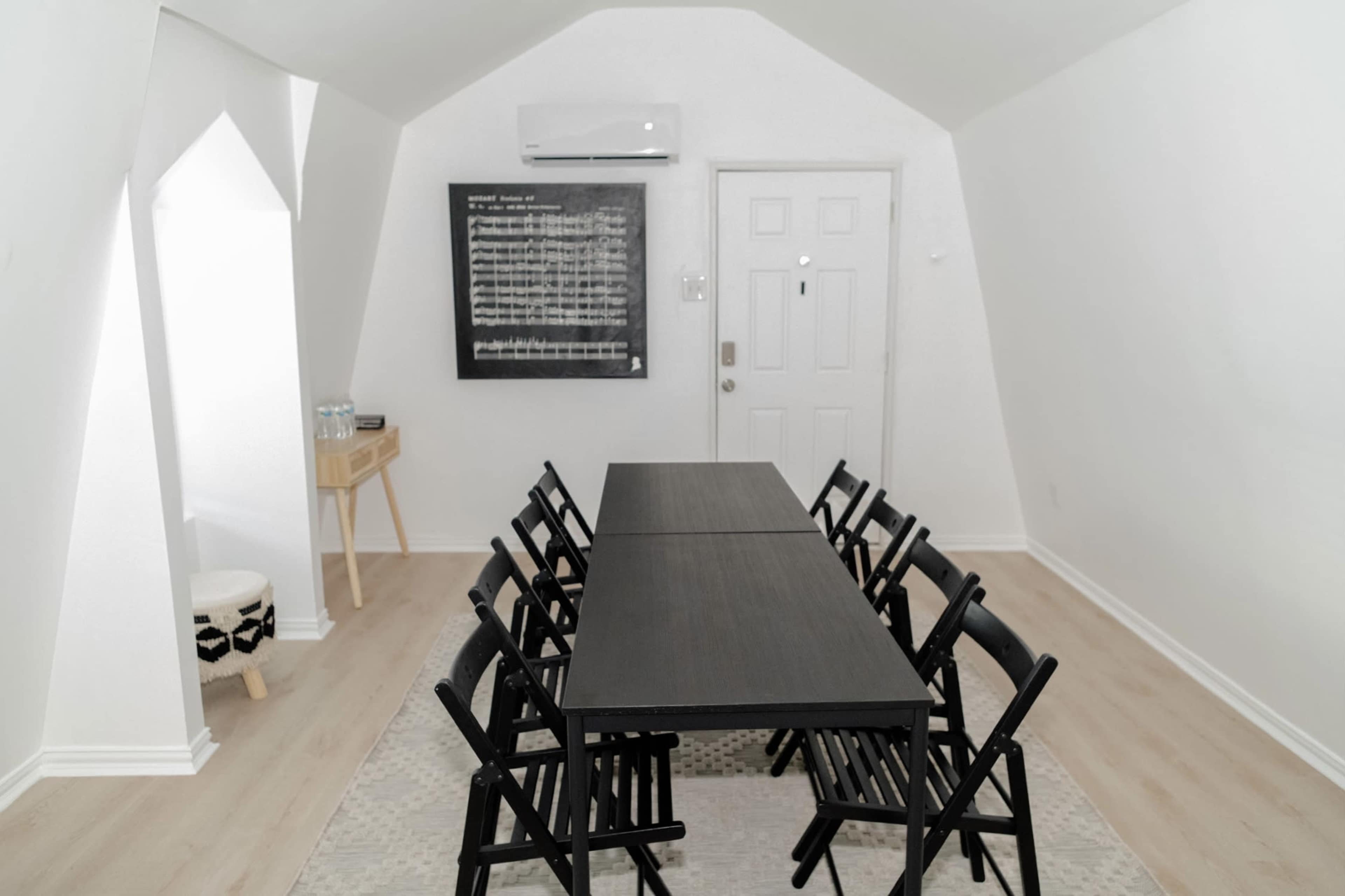 A minimalistic meeting room with a long black table surrounded by eight folding chairs, a wall-mounted air conditioner, and a large blackboard on the wall.