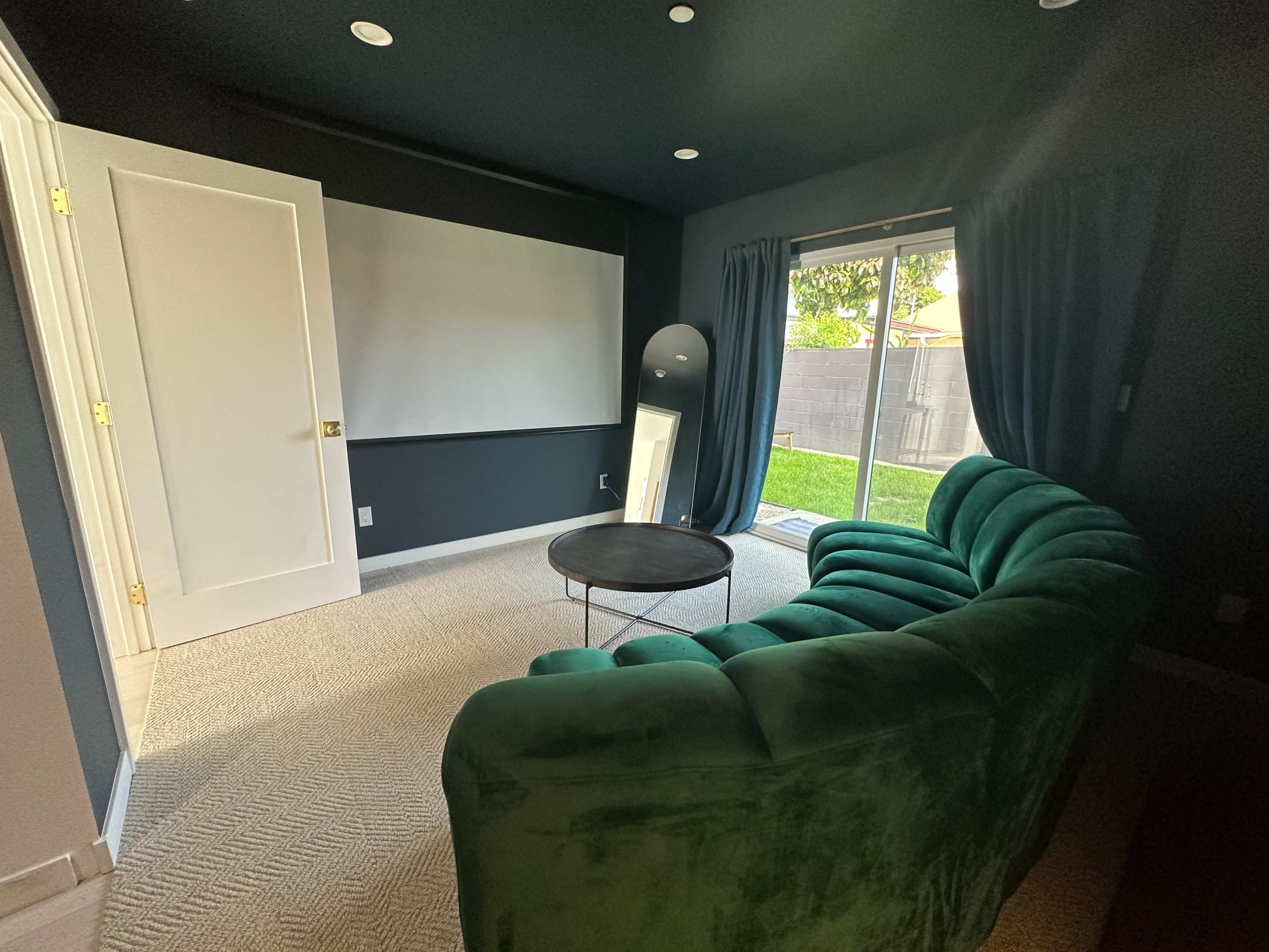 The room features a green velvet sofa, a round coffee table, a blank wall for projection, and large windows with curtains looking out to a grassy area.
