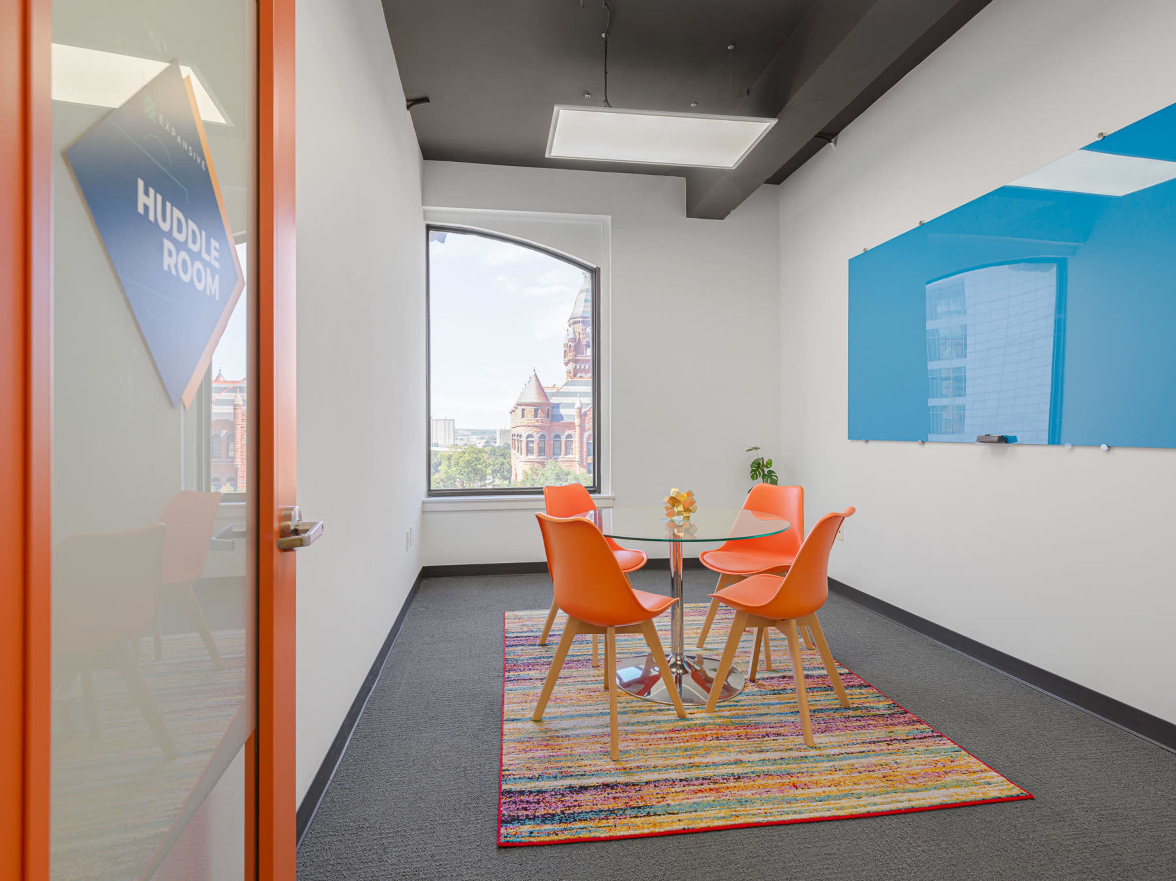 The image shows a small conference room with a round table and four orange chairs, featuring a large window and a blue whiteboard on one wall.
