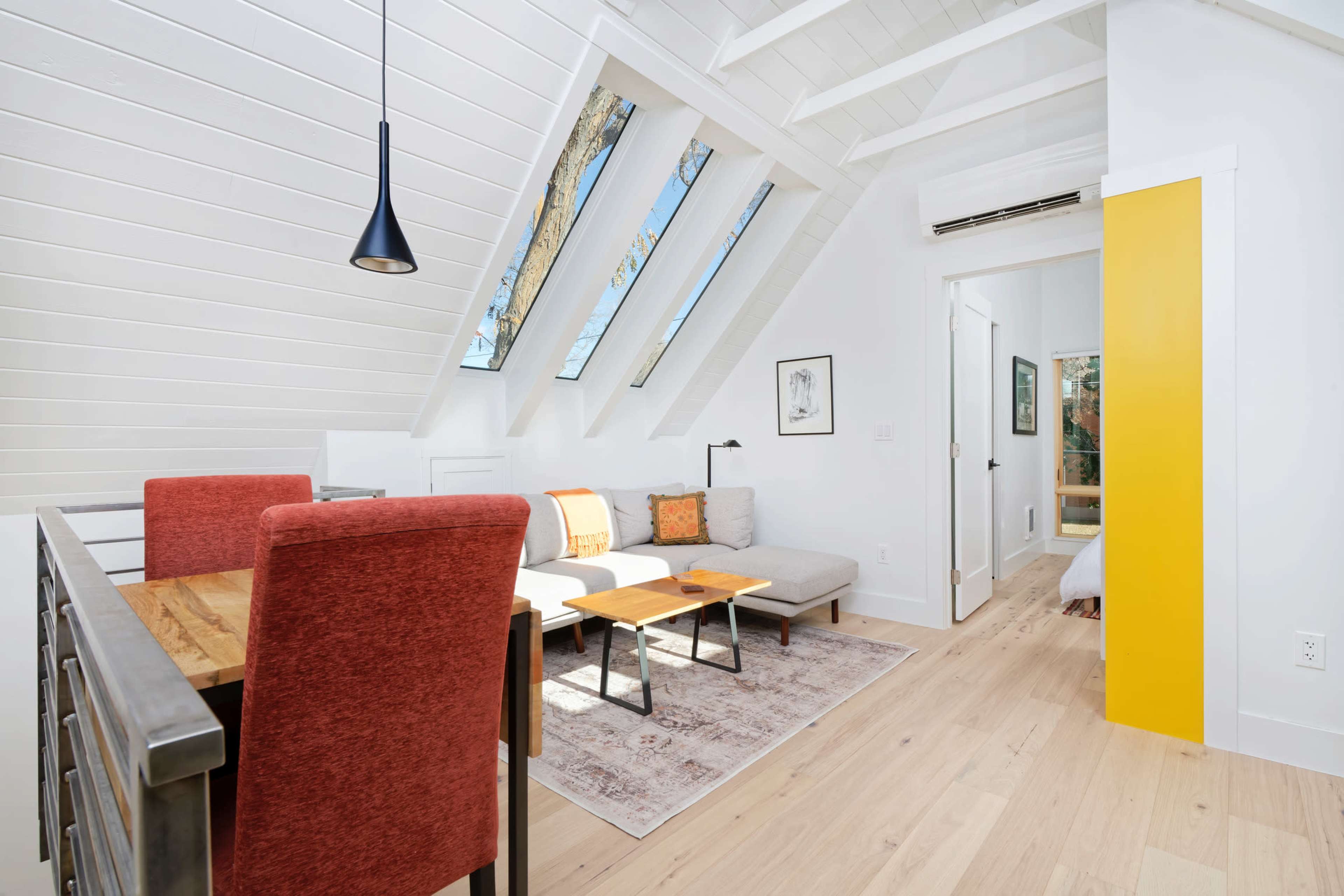 The image shows a bright, modern living space with sloped ceiling, large skylights, a dining area with red chairs, a gray sofa, and light wood flooring.