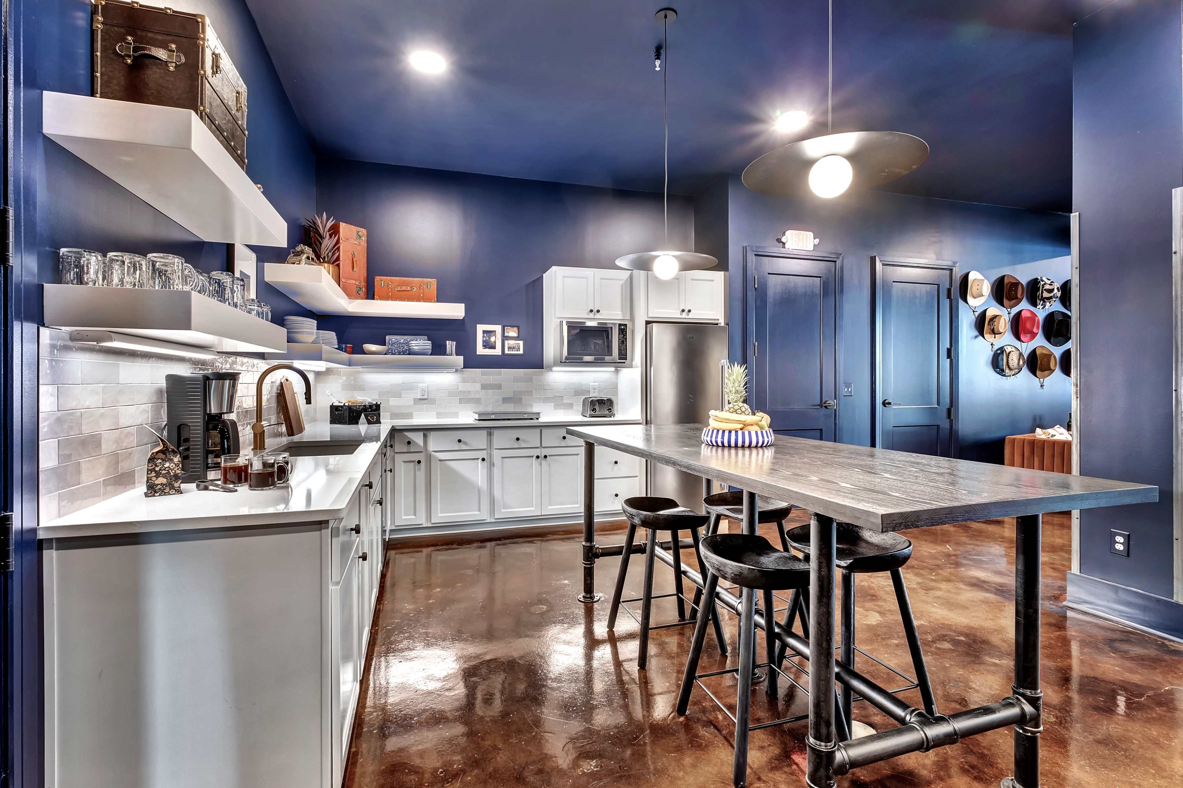 The image showcases a modern kitchen with dark blue walls, a central island with bar stools, white shelving, and stainless steel appliances.