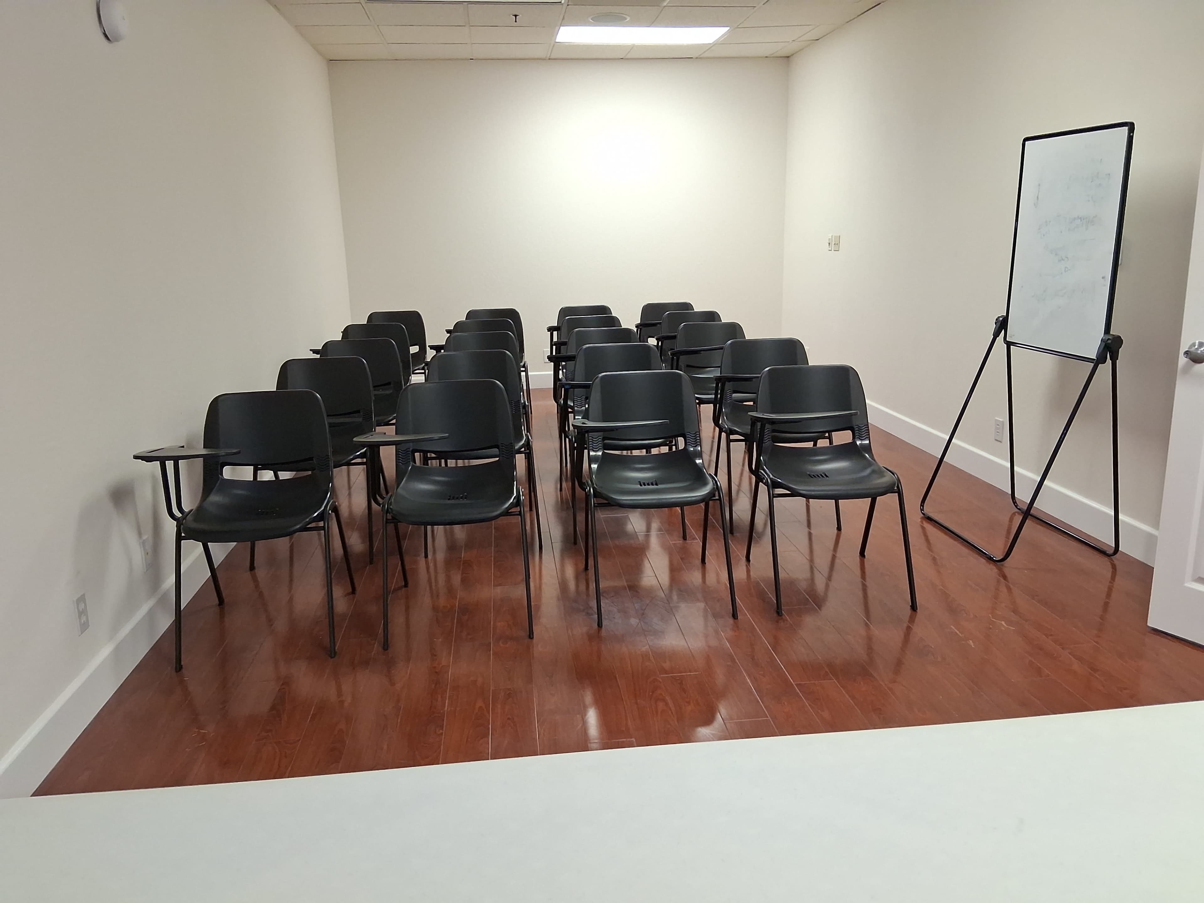 The image shows a sparsely furnished meeting room with rows of black chairs and a whiteboard at the front.