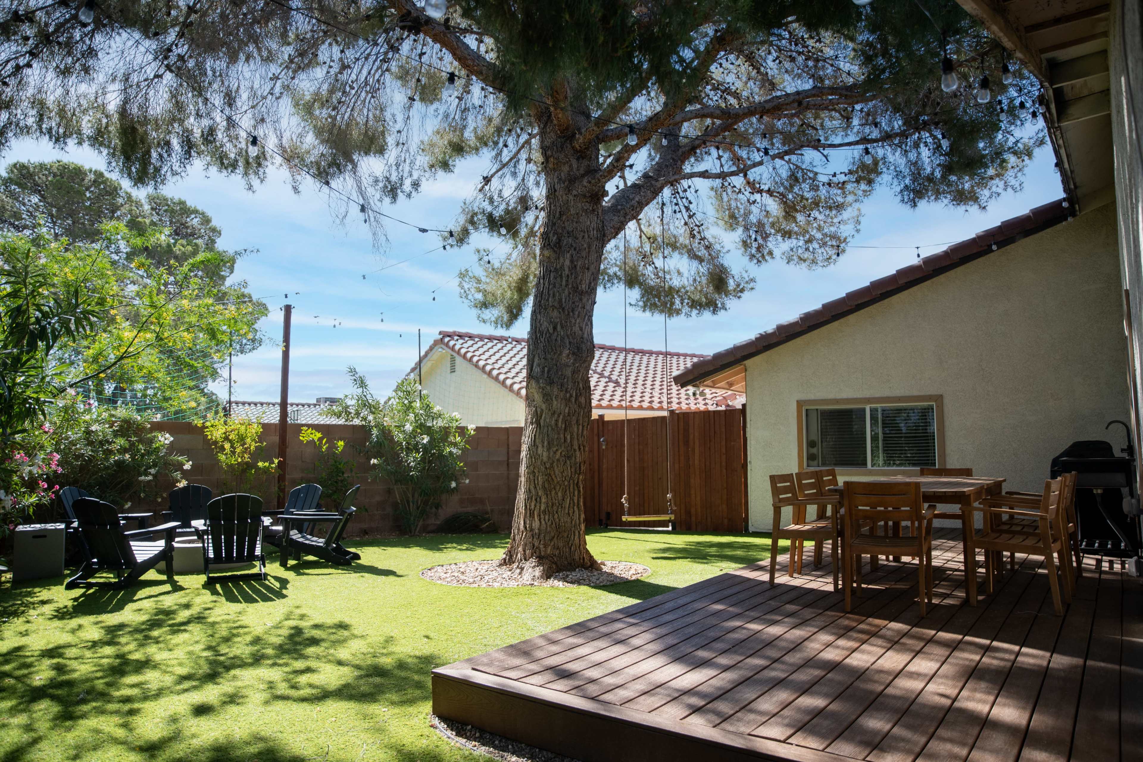The image shows a backyard with a wooden deck, a dining table, and several lounge chairs under a large tree, surrounded by landscaped grass and plants.