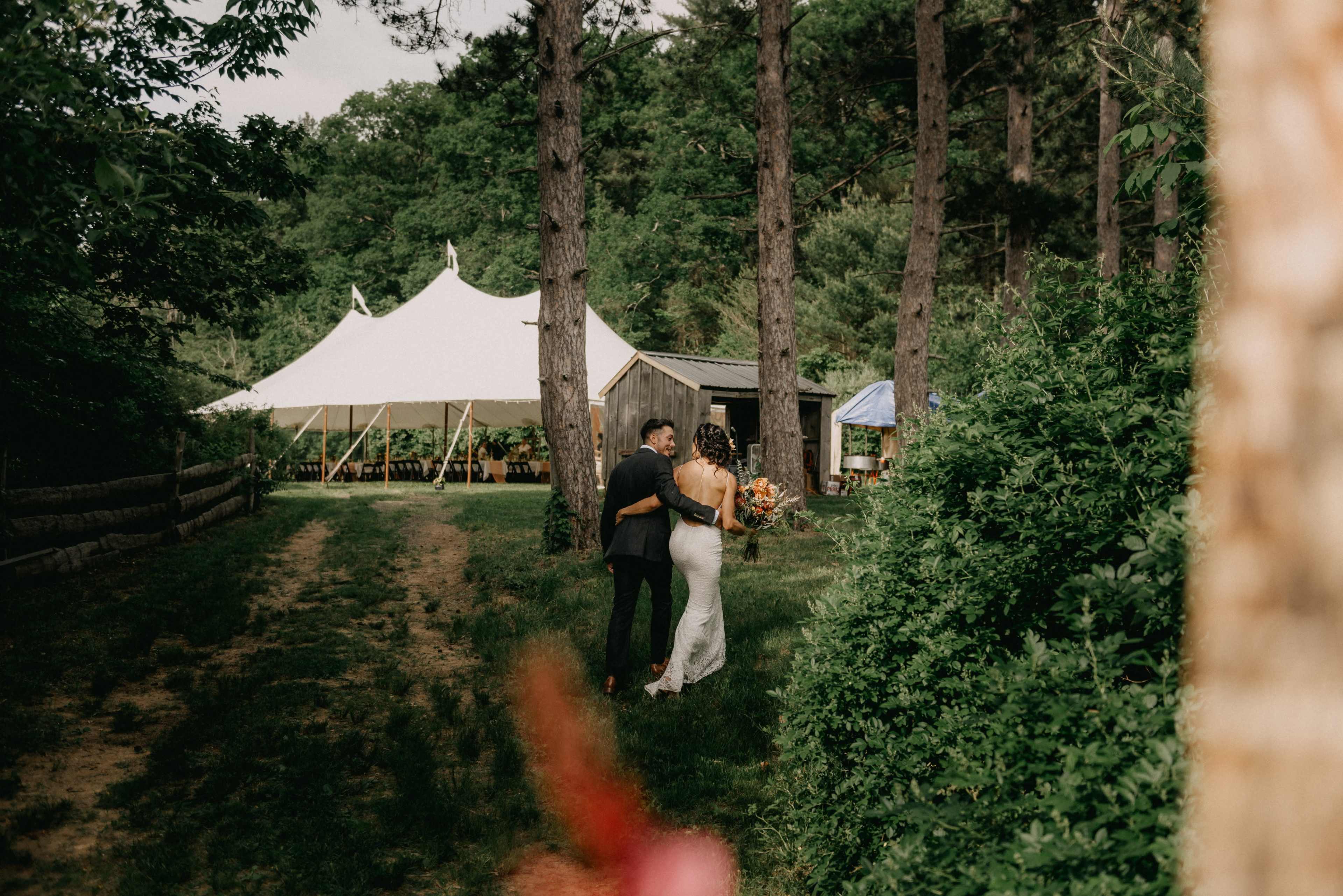 A couple embraces while walking along a grassy path leading to a large white tent set among trees.