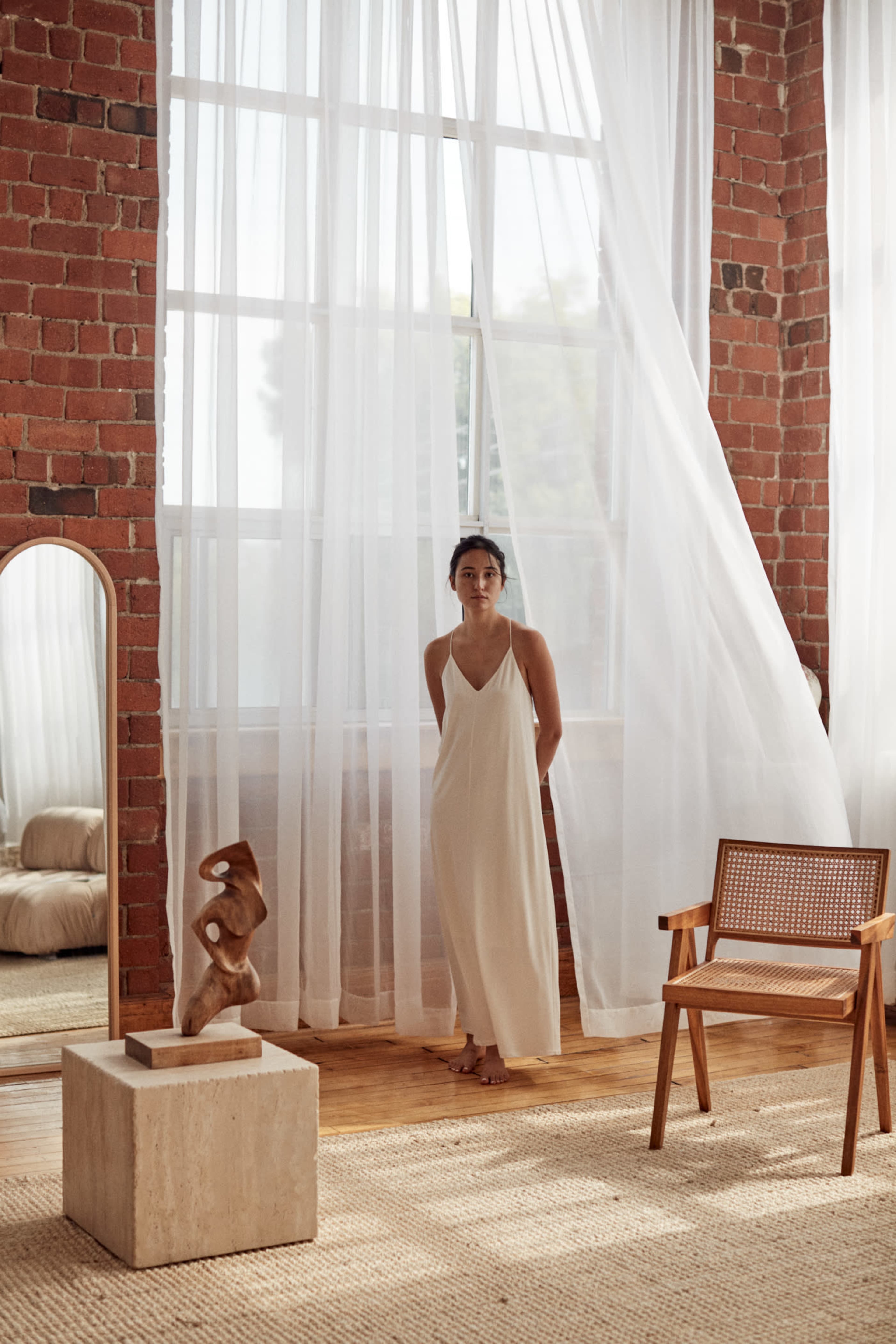 A woman stands by large windows draped with sheer curtains in a room featuring exposed brick walls and minimalist decor.