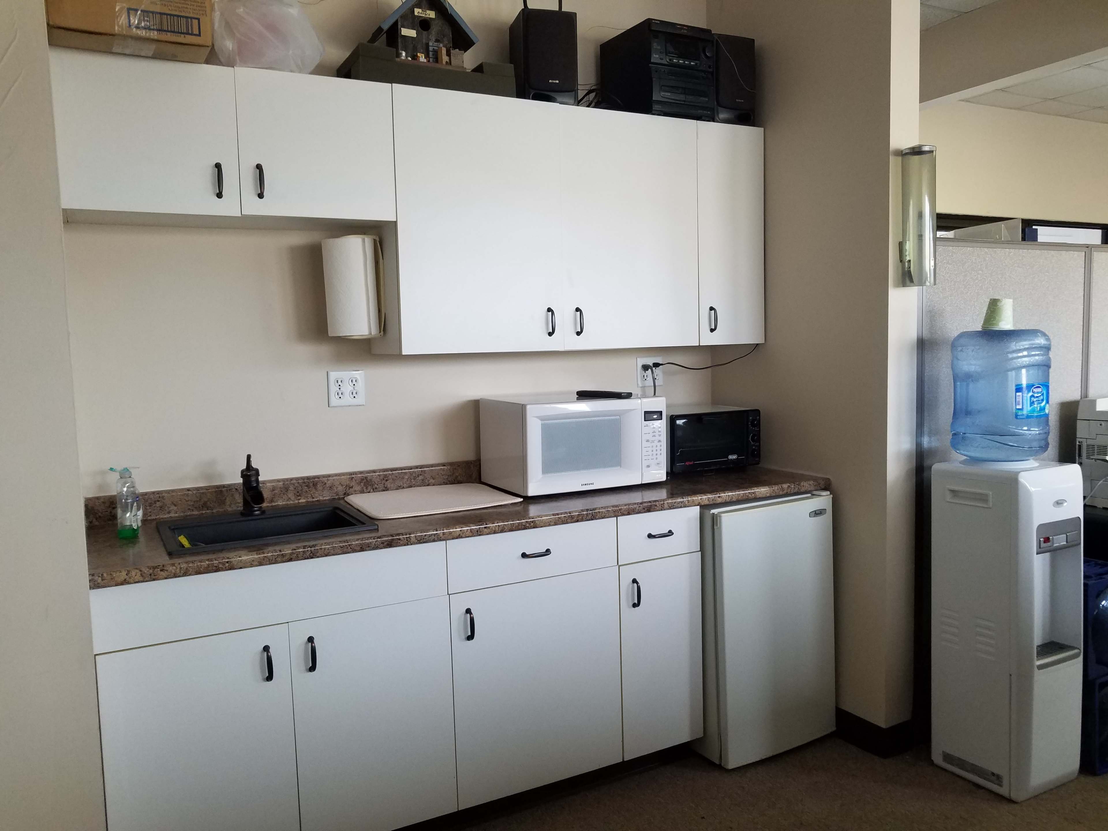 A kitchenette with white cabinets, a sink, a microwave, a small refrigerator, and a water cooler.