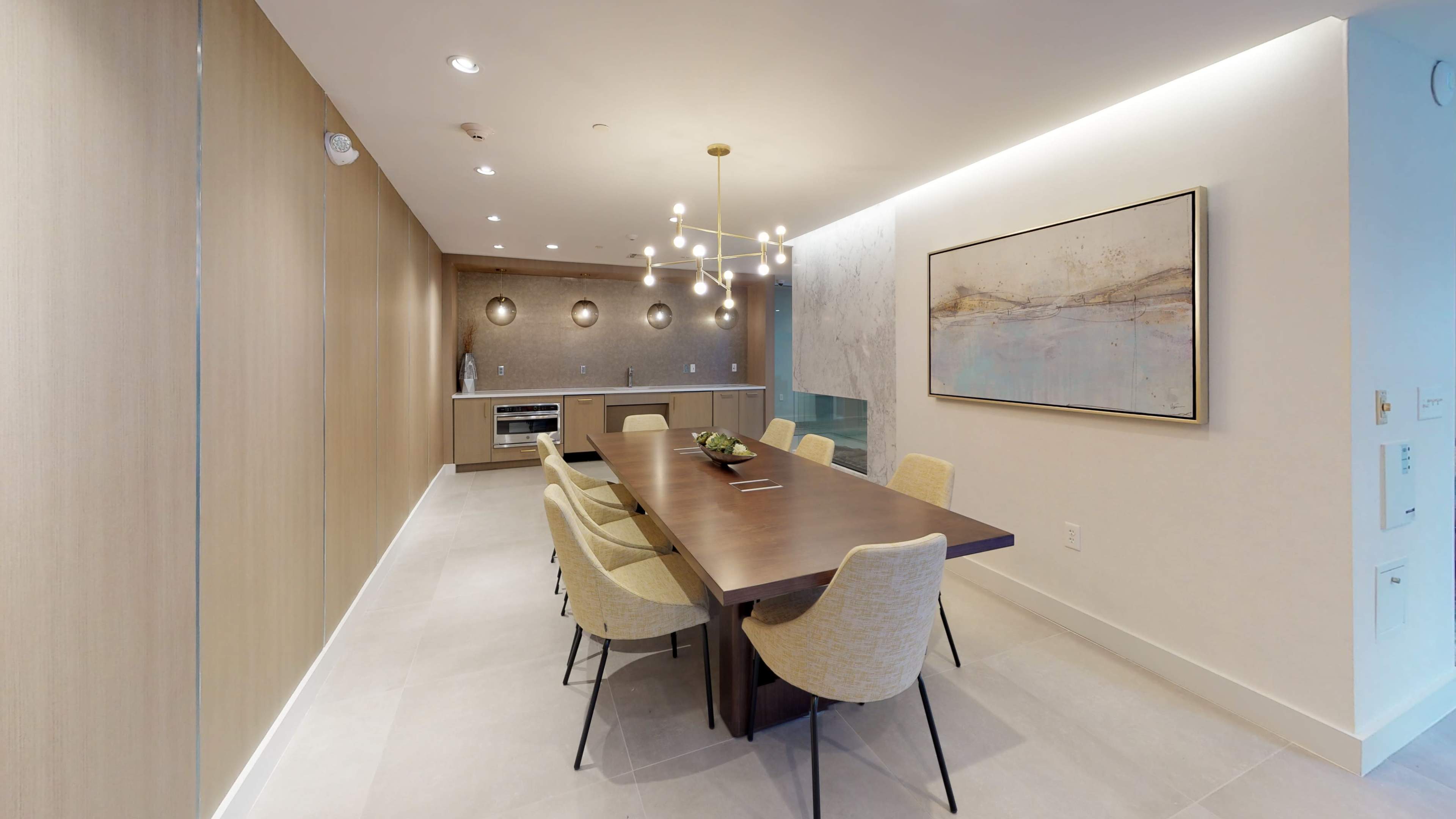 The image shows a modern dining room featuring a long wooden table surrounded by beige chairs, with a wall-mounted art piece and a kitchenette in the background.