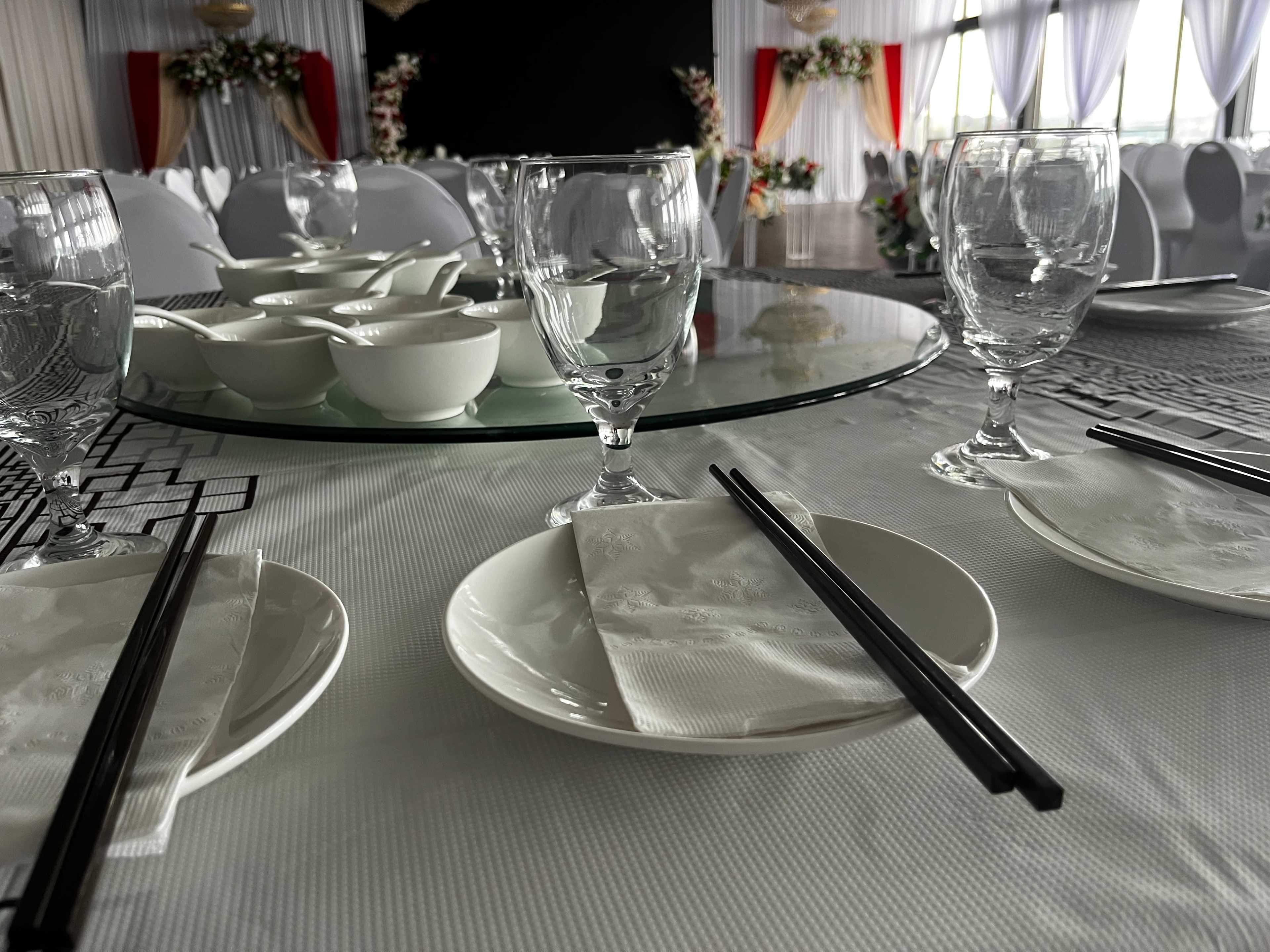 The image shows a dining setup with glassware, plates, chopsticks, and a circular glass table surrounded by chairs in a decorated space.