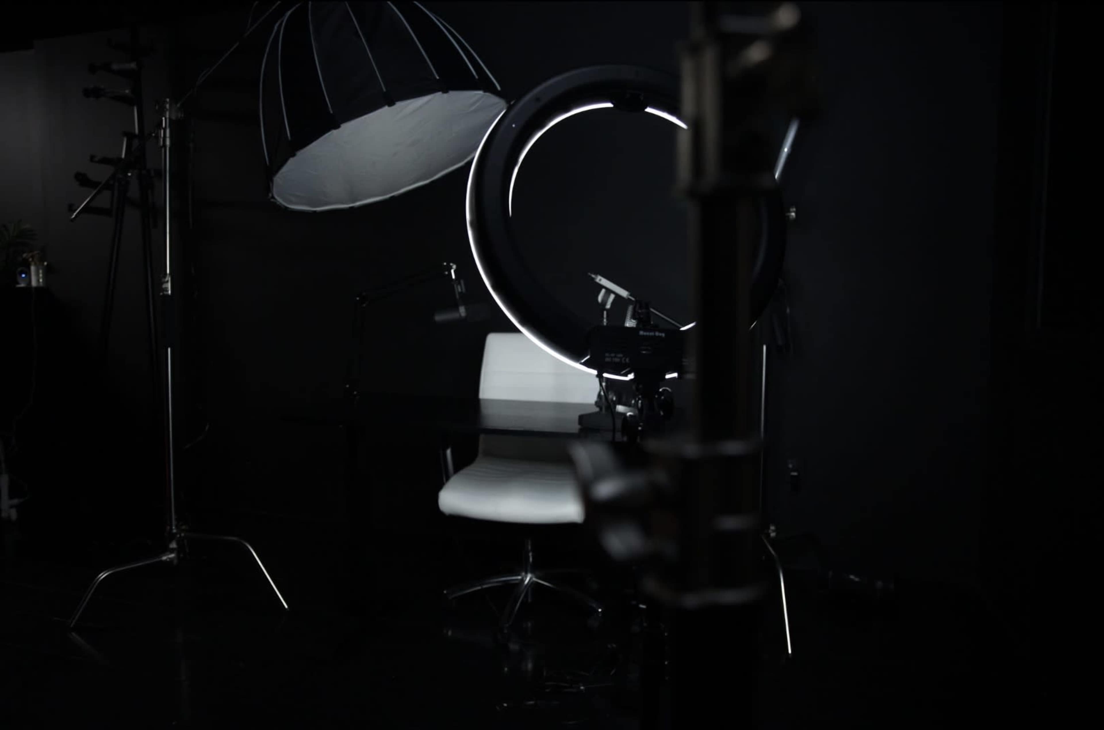 A photography studio setup featuring a circular LED light, a softbox, and a white chair against a dark background.