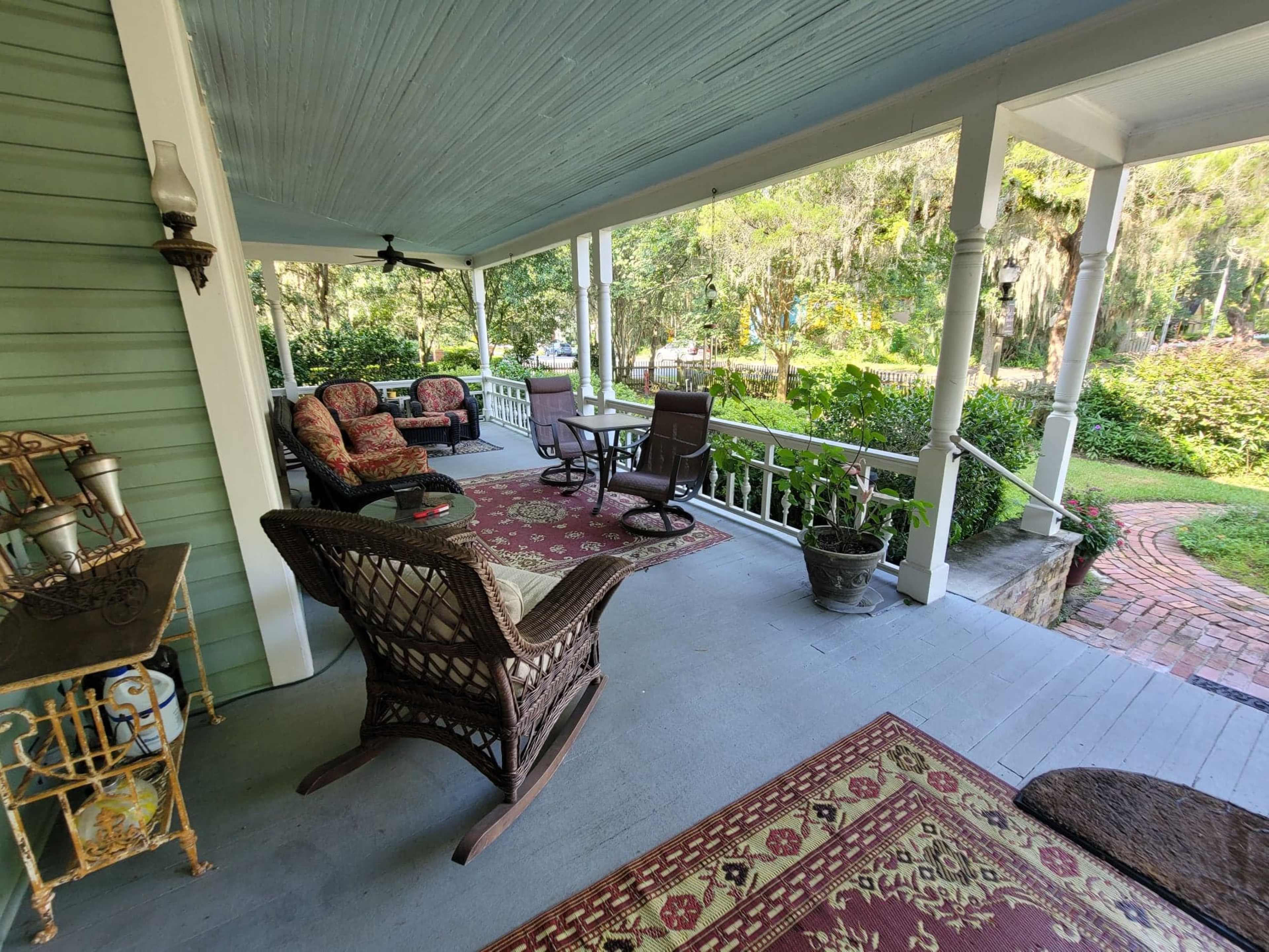 A covered porch with seating arrangements, including wicker chairs and cushions, overlooking a garden.