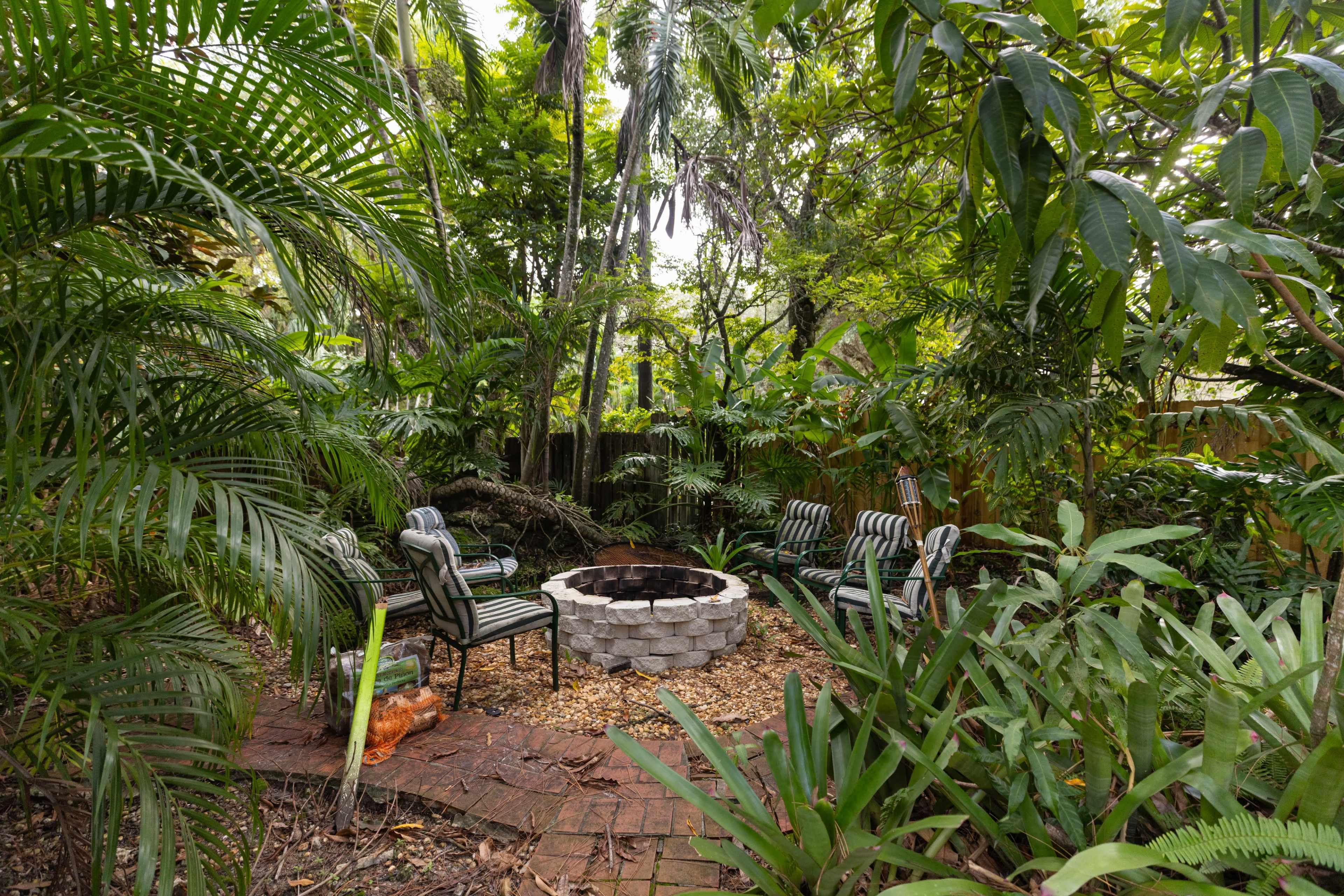 The image shows a circular stone fire pit surrounded by four chairs in a densely vegetated backyard.