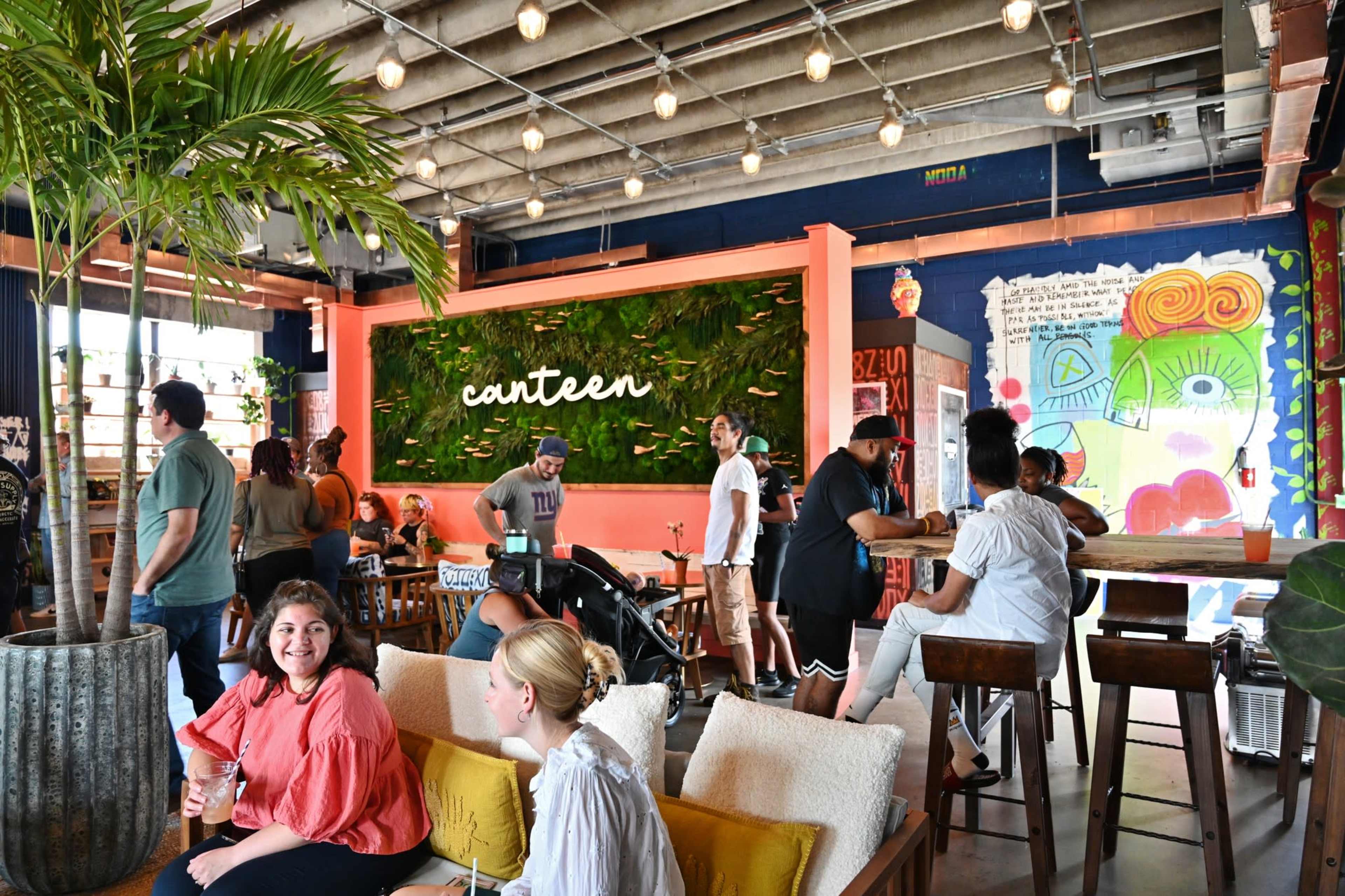 A bustling café interior features patrons socializing, a large green wall with the word "canteen," and vibrant mural art.