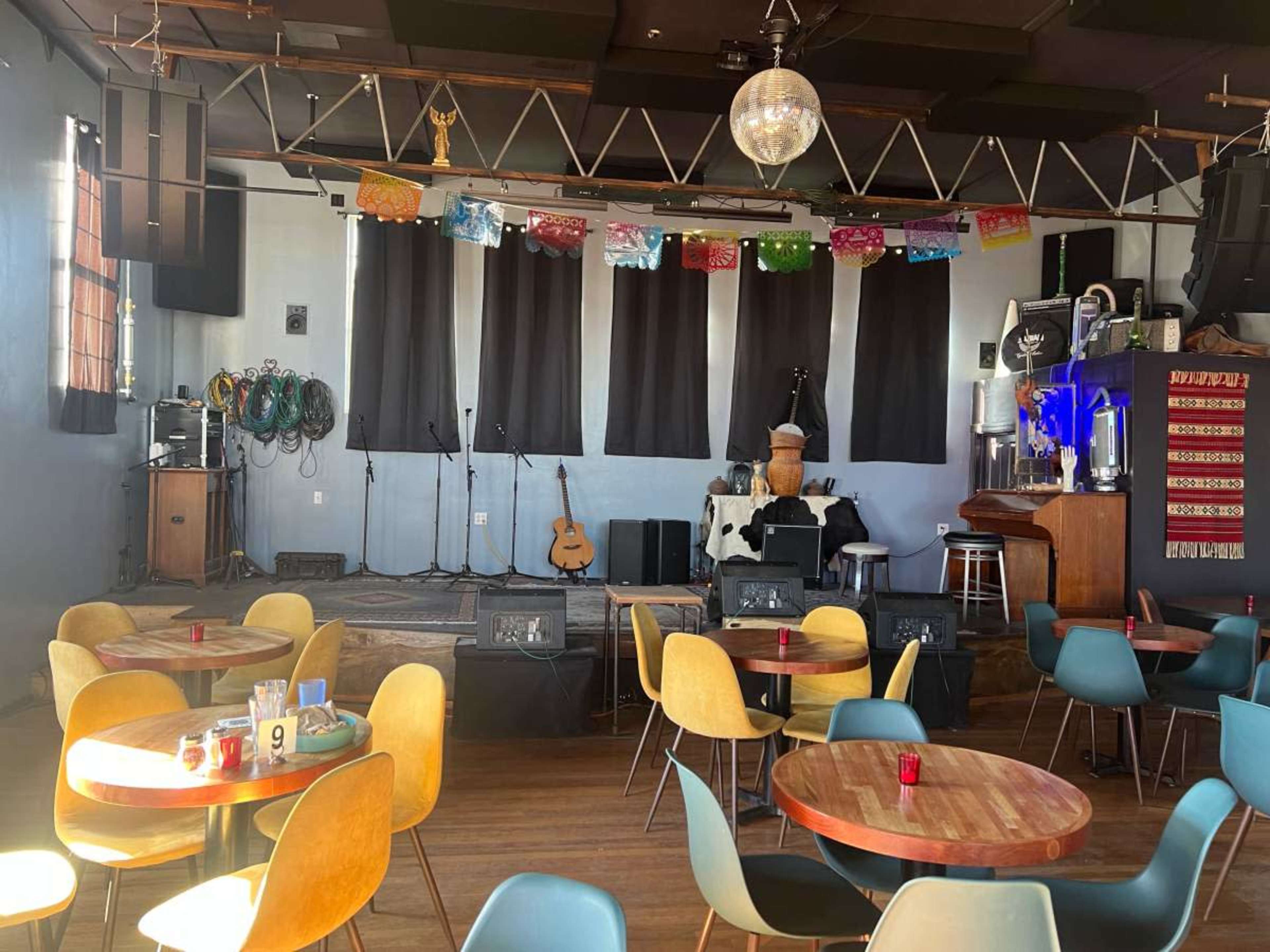 The image shows a colorful music venue with tables and chairs arranged around a small stage featuring a guitar and various musical equipment, along with decorative banners hanging from the ceiling.