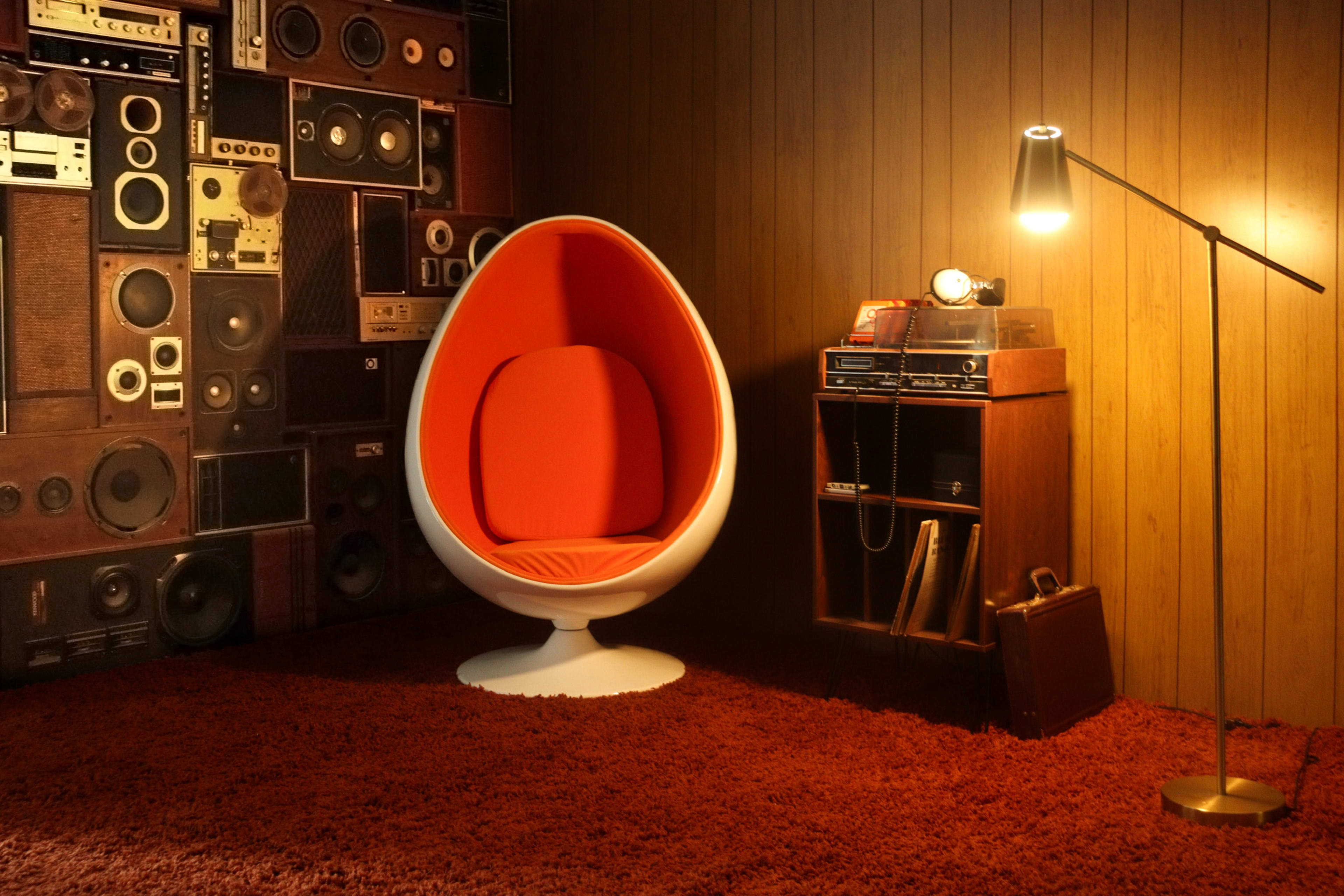 The image shows a retro-themed room featuring an orange egg-shaped chair, a wooden cabinet with audio equipment, and a floor lamp, all set against a wall adorned with vintage speakers.