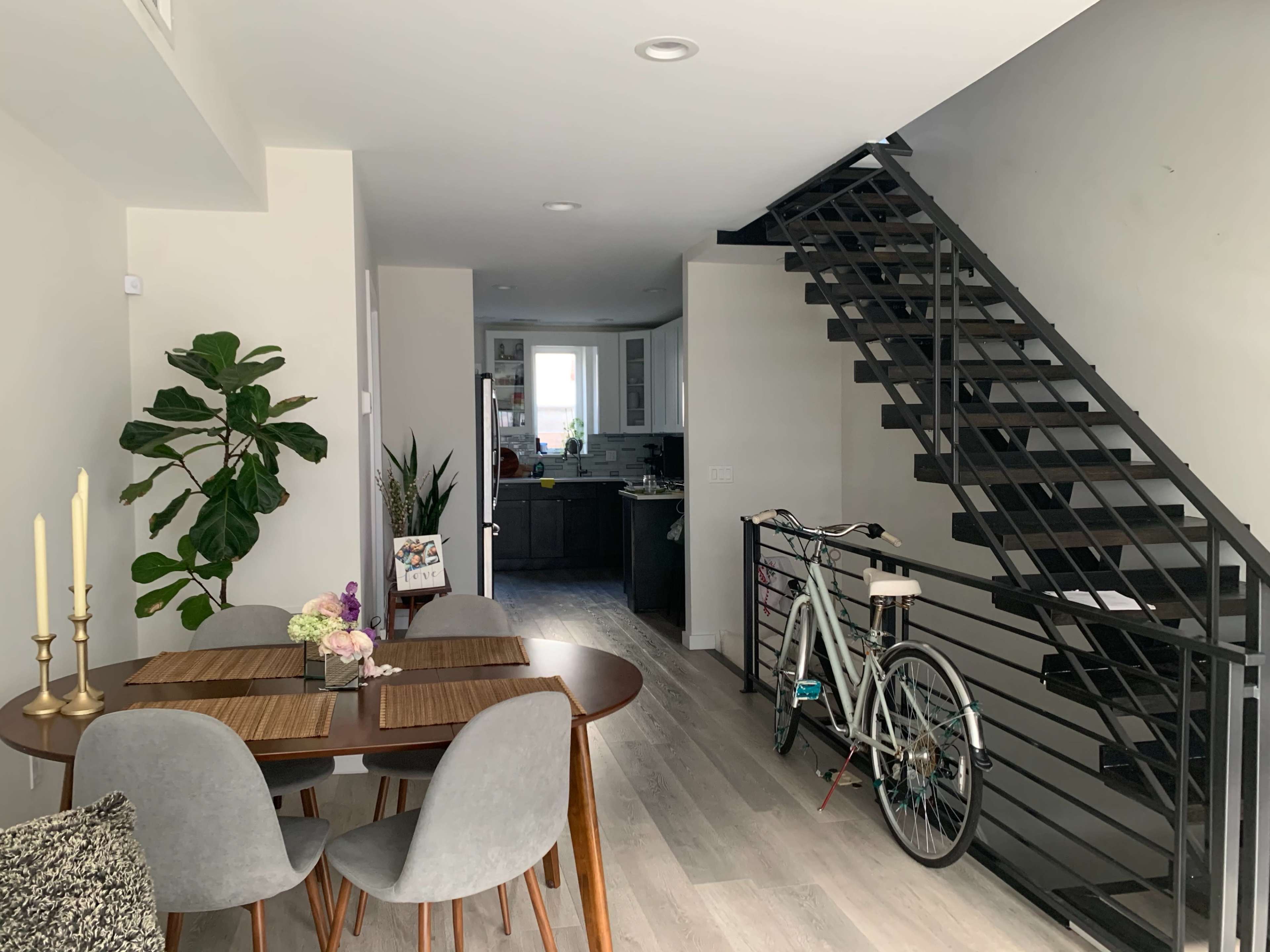 The scene shows a modern interior with a dining area featuring a round table and chairs, a plant in the corner, a staircase, and a bicycle parked against the wall near a doorway leading to a kitchen.