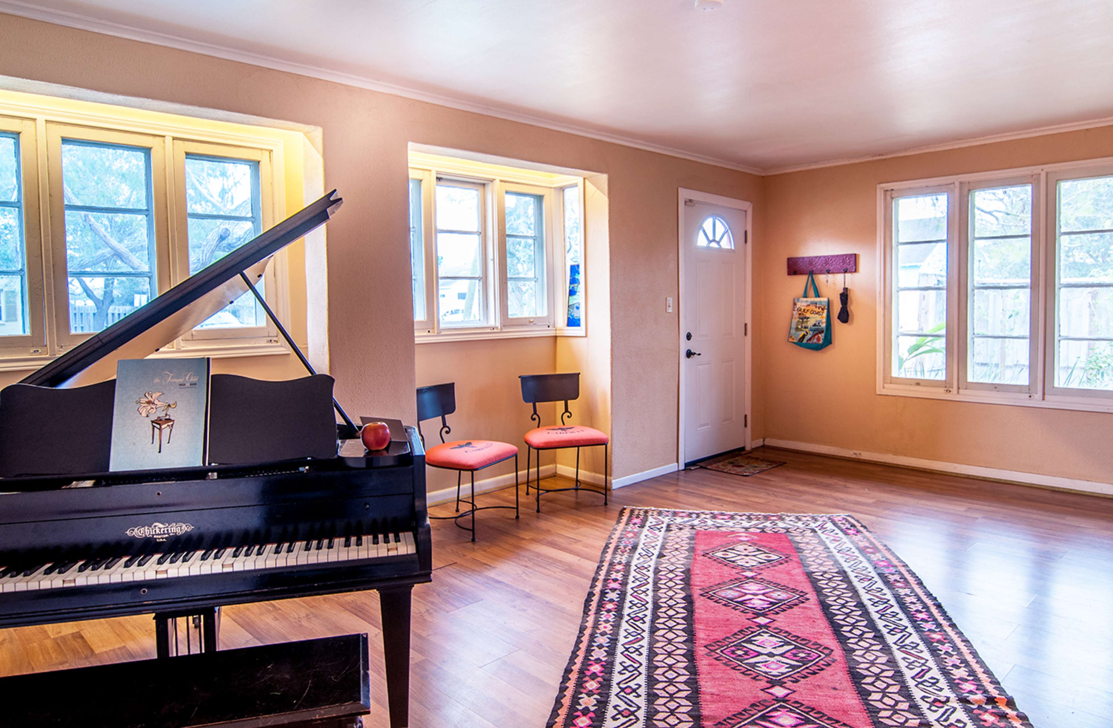 The room features a grand piano on the left, two chairs with red cushions, a patterned rug on the wooden floor, and several windows illuminating the space.