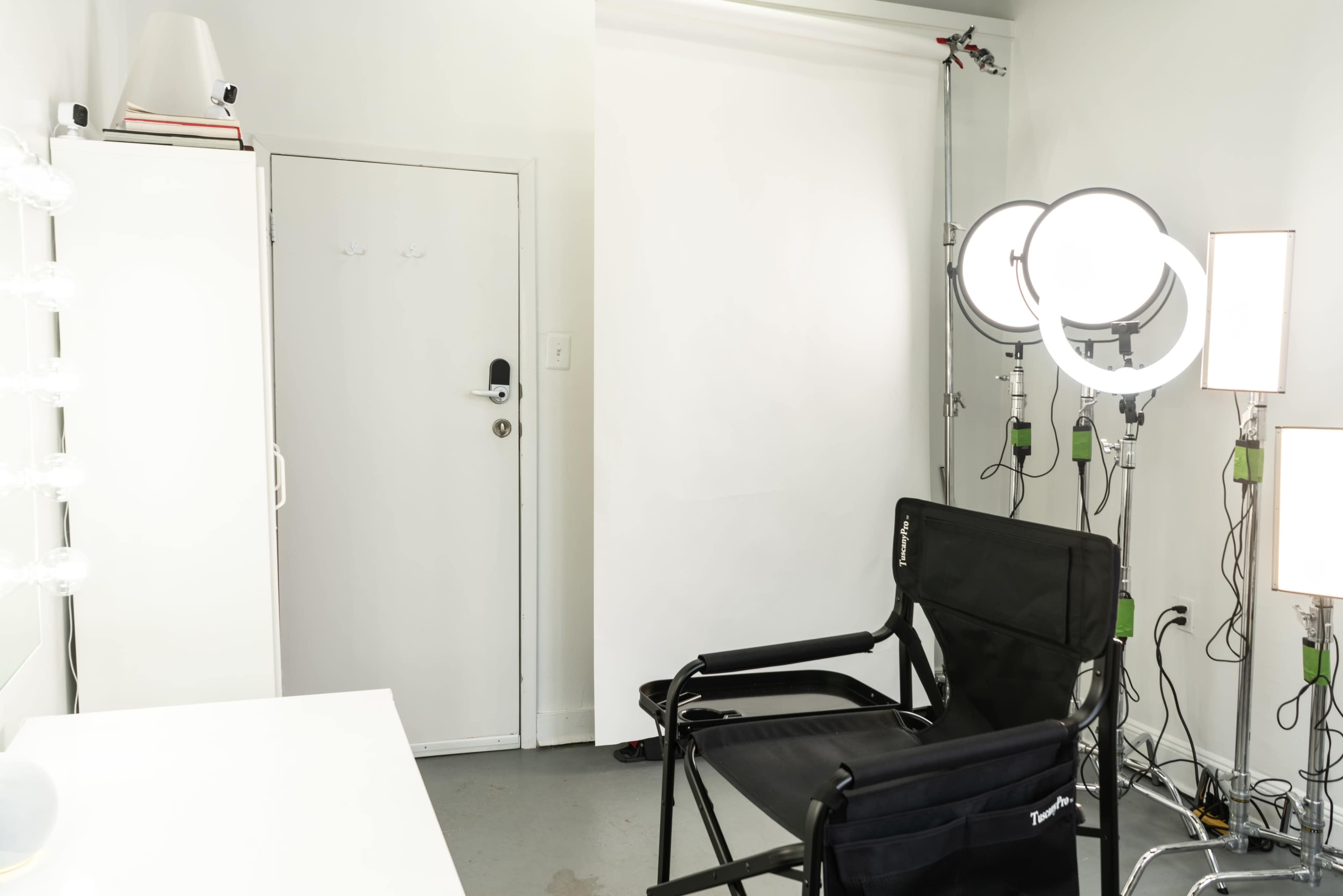 A makeup studio features a black director's chair, a white backdrop, and multiple light fixtures.