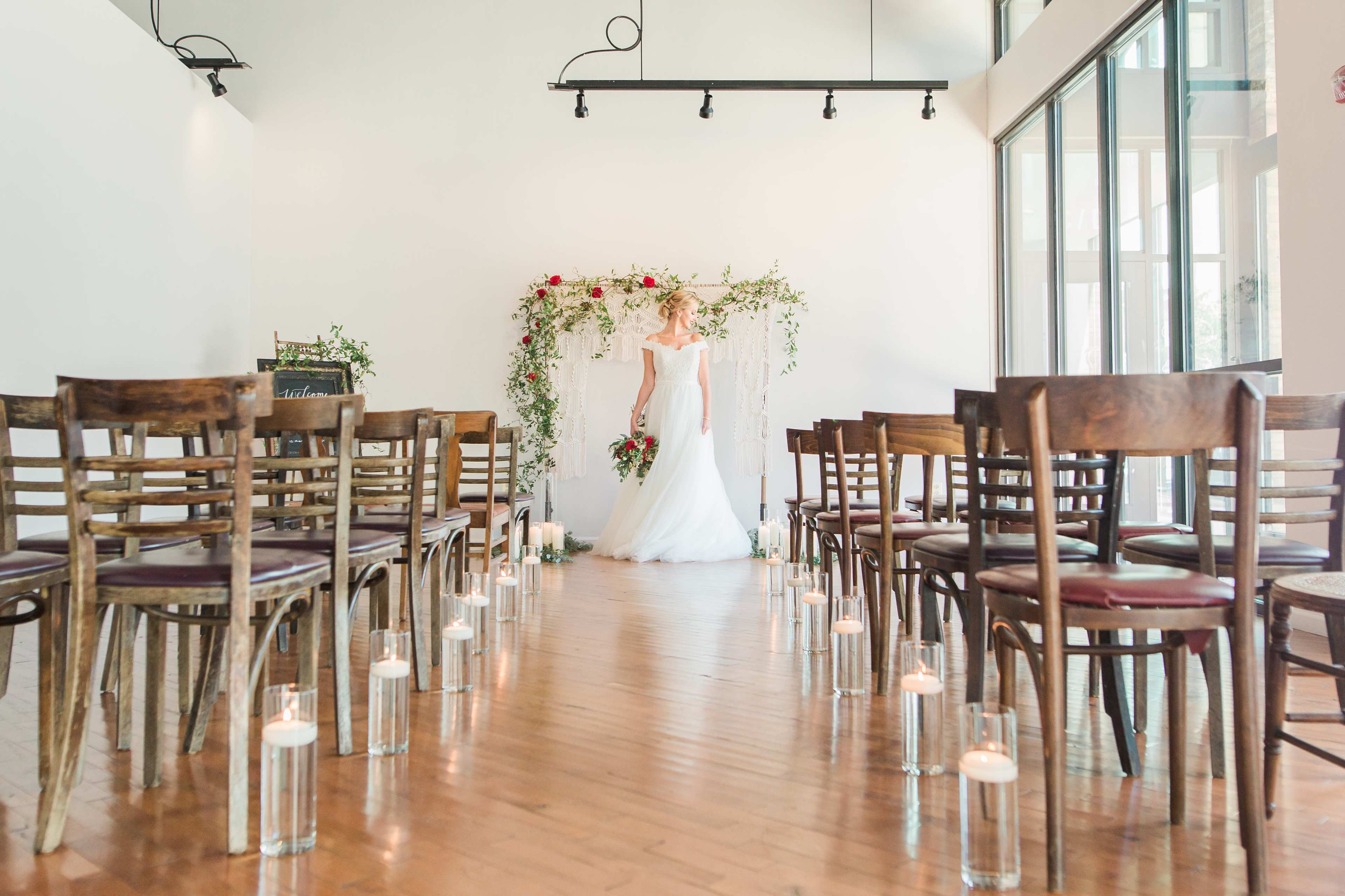 A bride stands in front of a floral archway at the end of an aisle lined with candles in a spacious, well-lit venue.