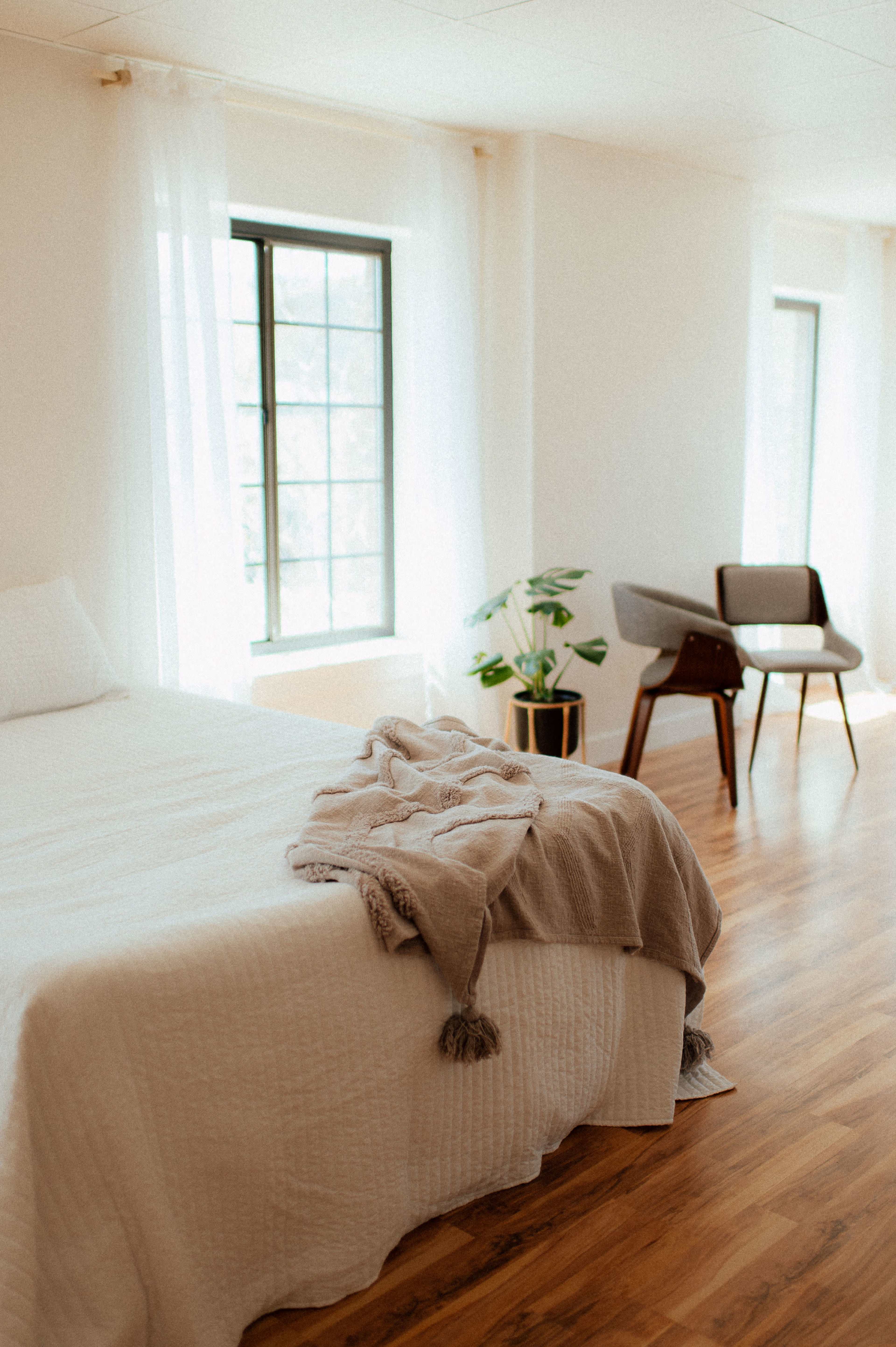 A bright bedroom features a bed with a light quilt, a plant in a pot, and a stylish chair near windows with sheer curtains.