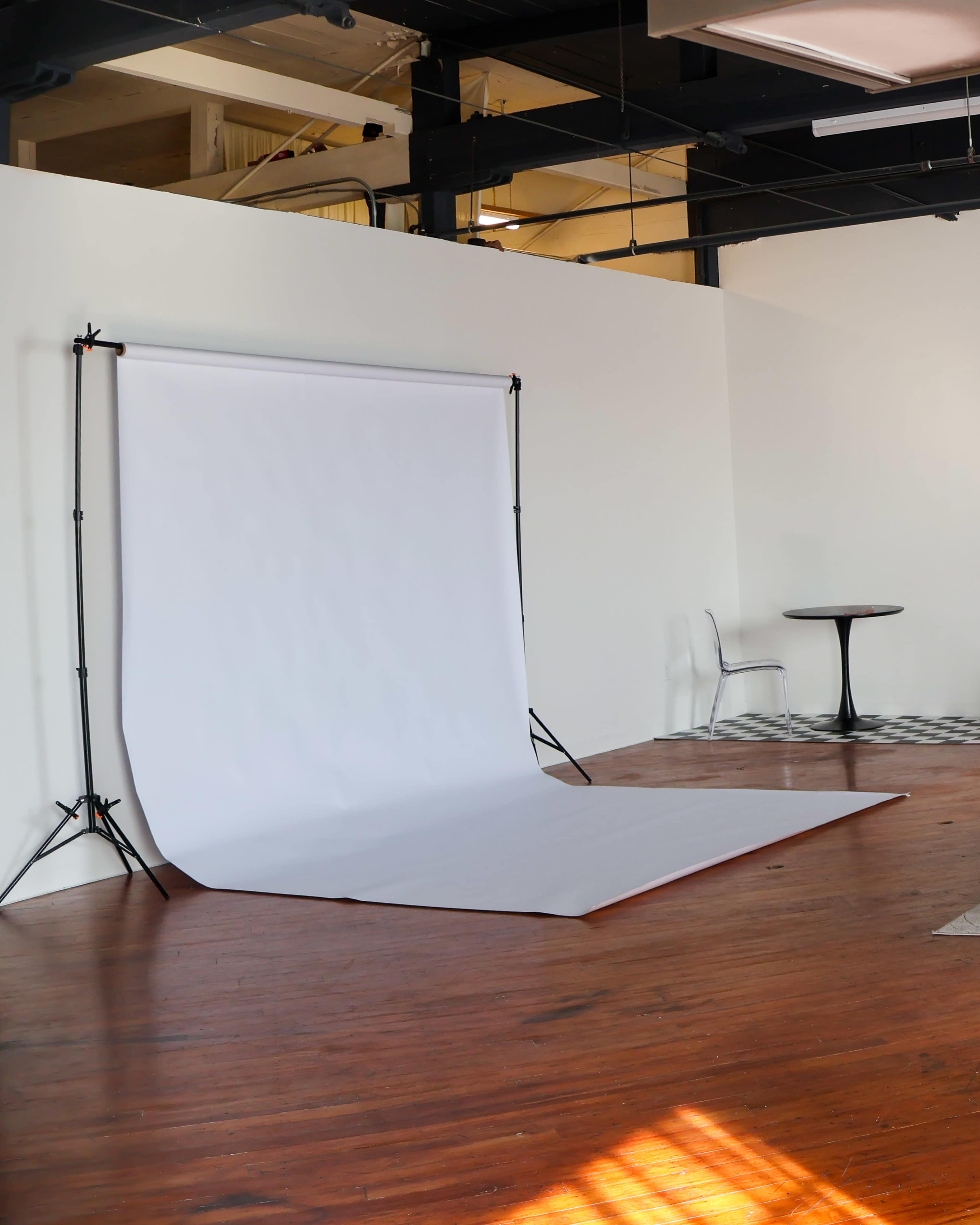 The image shows a photography studio with a white backdrop set up on a wooden floor, accompanied by a small table and a chair in the corner.
