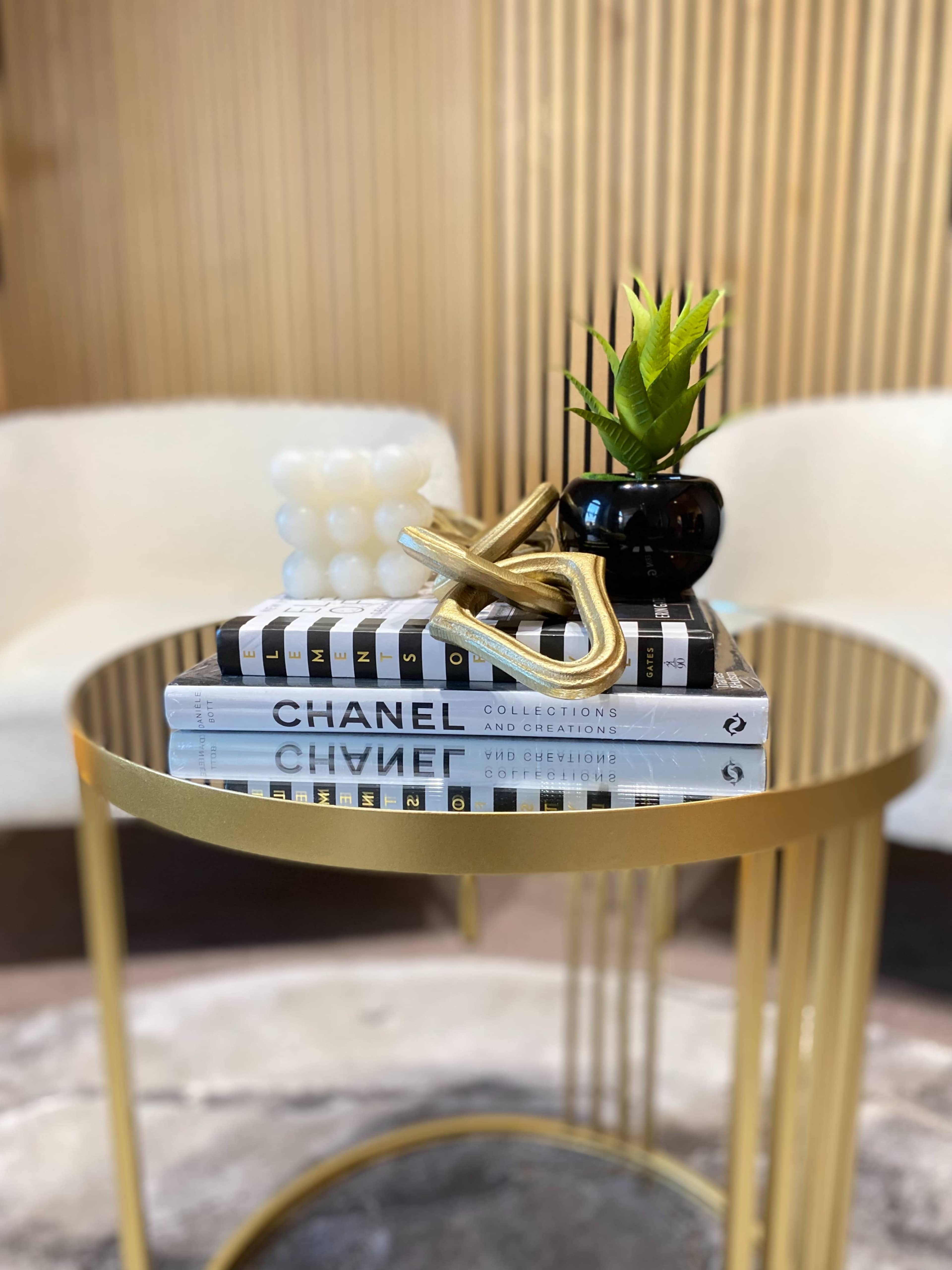 A round glass table displays stacked books and a decorative gold sculpture, accompanied by a small black vase holding a green plant, all set against a backdrop of wooden panels.